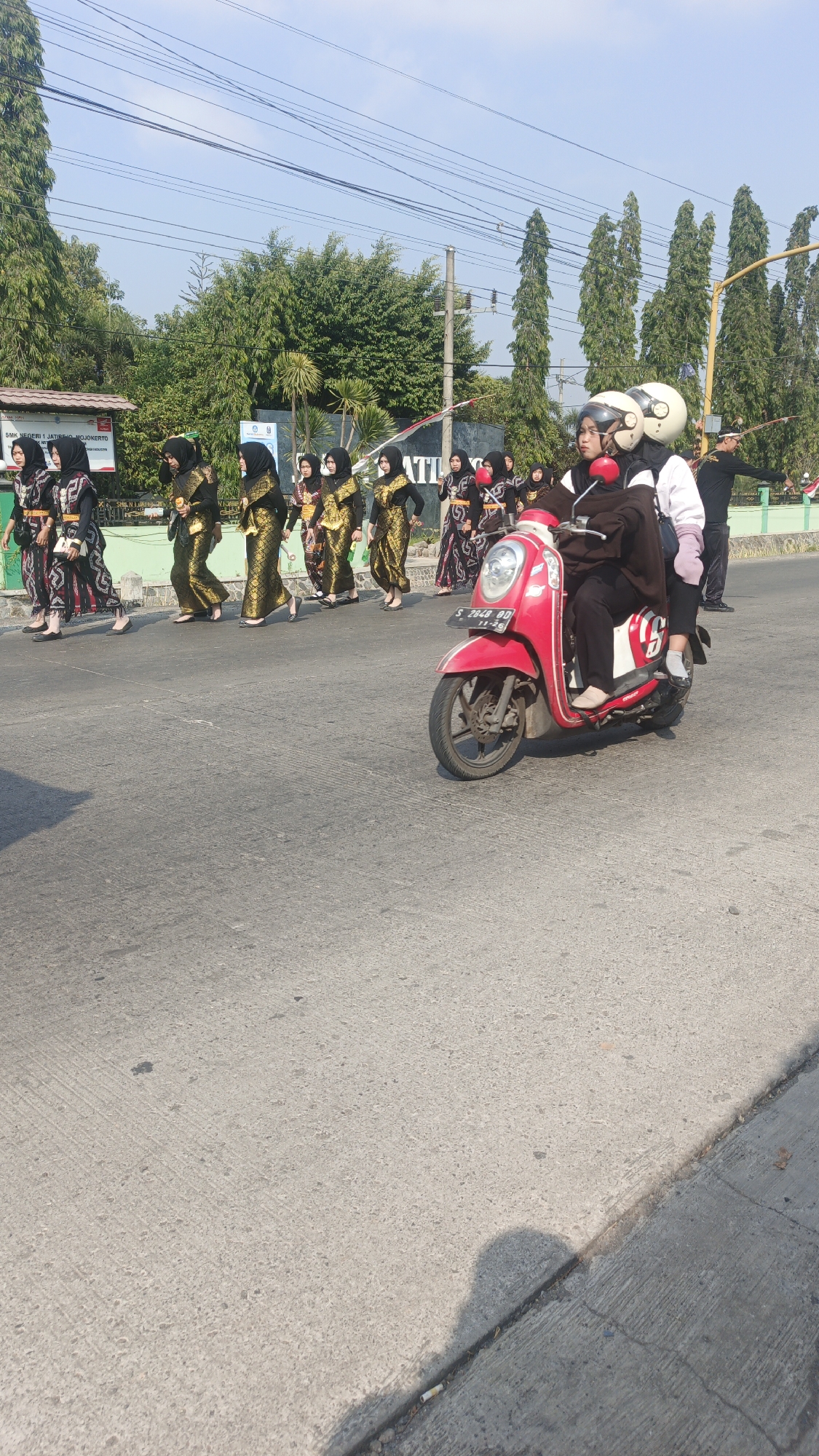In Sumengko, Indonesia, on a paved road under a clear sky, a group of people dressed in traditional attire, likely for a parade or cultural event, walk in single file. They are adorned in black and gold patterned clothing and headscarves, suggesting a formal occasion. To the right of the procession, a red scooter carries two individuals wearing helmets. The rider, wearing a dark brown outfit, navigates the scooter, while a passenger in a white top and pink pants sits behind. In the background, a building with signage that reads "SMK NEGERI 1 JATIBONGOOERTO" and "ATI" is visible, along with tall trees and utility poles, indicating an urban or suburban setting. The lighting suggests it is daytime, with shadows cast on the road, indicating a sunny environment.