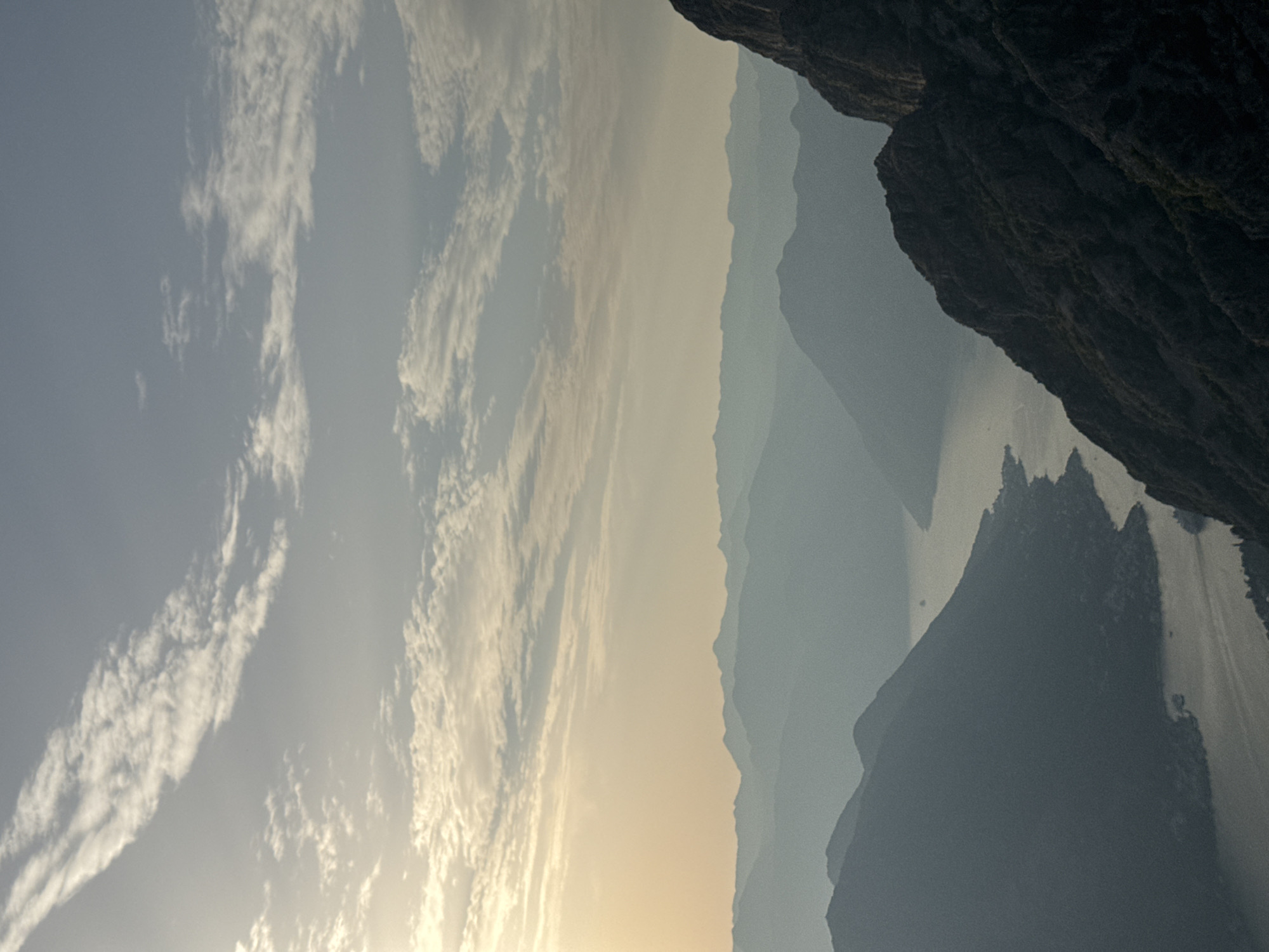 The image captures a wide, scenic view of a bay with surrounding mountains, likely from a high vantage point. The sky is filled with wispy clouds, and the sun is low on the horizon, casting a soft, warm light that creates a hazy, atmospheric effect over the distant mountain ranges. The foreground features a dark, rocky mountain slope on the right, leading the eye into the scene. In the middle ground, a body of water, possibly a bay or a fjord, winds between layers of silhouetted mountains that recede into the distance, becoming progressively lighter and fainter due to atmospheric perspective. A few small boats are visible on the water. The environment suggests a coastal mountainous region. The location is specified as Žanjev Do, Montenegro, which is known for its dramatic coastal landscapes. The image does not contain any people or discernible actions.