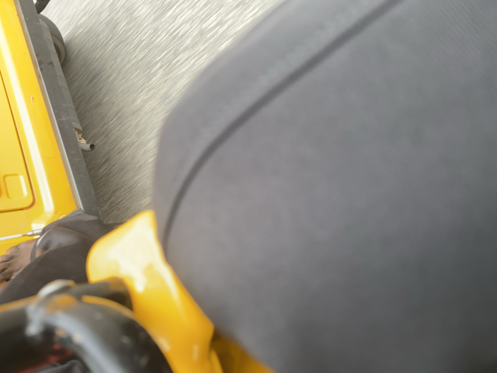The image is a first-person perspective shot from what appears to be the driver's seat of a yellow motorized rickshaw, likely an auto-rickshaw or "Keke Napep", common in Nigeria. The foreground is dominated by a large, dark gray upholstered seat, suggesting the passenger seat or a portion of the driver's seat. To the left, the bright yellow body of the rickshaw is visible, with a glimpse of a black metal handlebar or frame in the lower left corner. A person's bare foot is visible on the floor of the rickshaw, near the yellow bodywork, with black pants also visible.
The background shows a blurred, streaky texture of gray and white, indicating motion, likely from the rickshaw moving along a road. The nature of the background suggests a paved road with some texture, possibly wet or dusty. The lighting appears to be bright, suggesting daytime, with no indication of specific weather conditions beyond the impression of movement. No visible text is present in the image. The scene is set in Jalingo, Nigeria, implying an urban or semi-urban environment where such vehicles are a primary mode of transport. The activity taking place is simply travel, with the perspective suggesting the person is either driving or a passenger in the rickshaw.