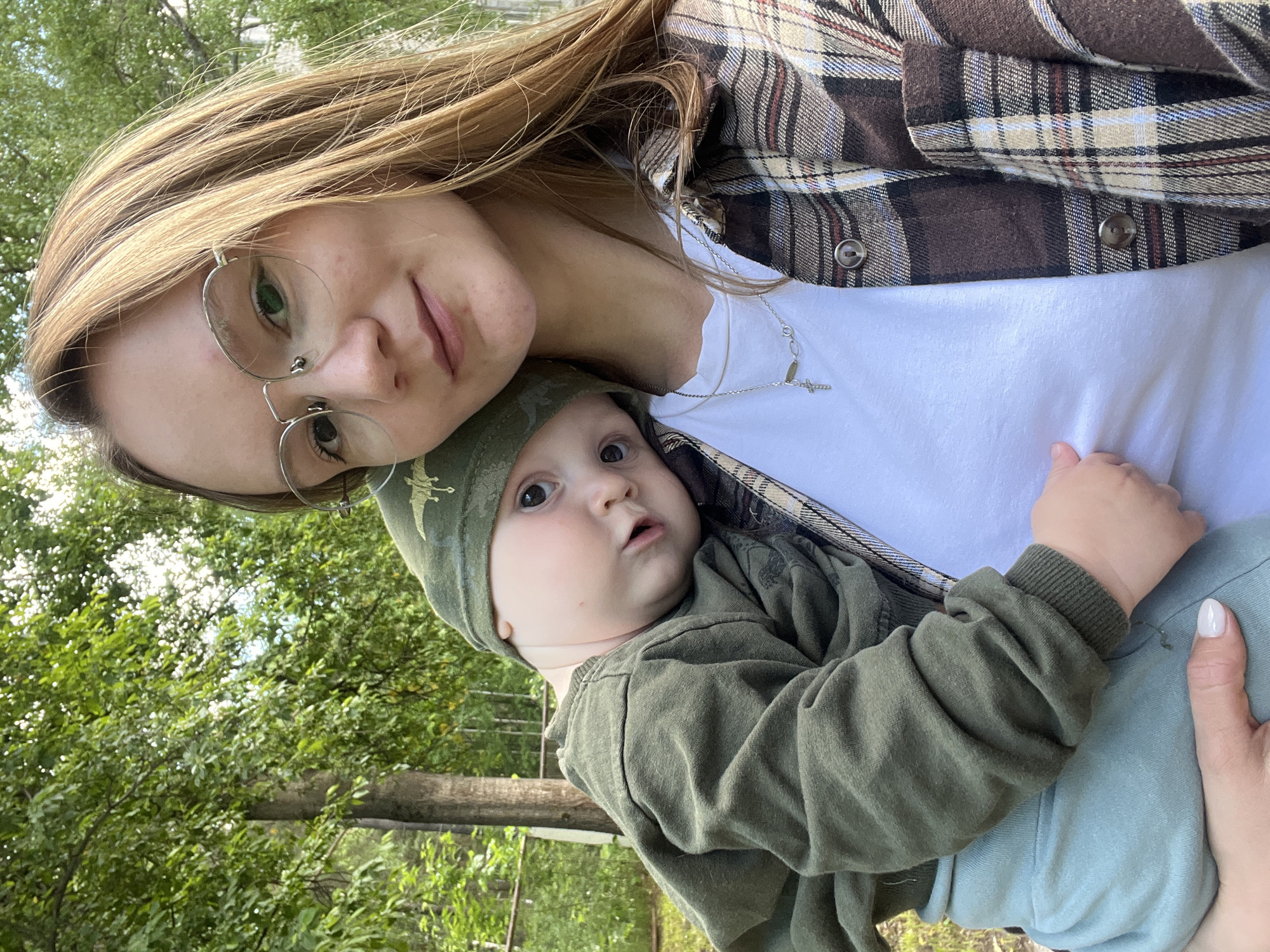A woman with light brown hair and glasses holds a baby outdoors. The woman wears a white t-shirt under a brown plaid shirt. The baby is dressed in an olive green outfit with a matching cap. The background shows green trees and vegetation, suggesting a park or wooded area in the city of Chaikovsky, Russia. The image is a selfie taken from a high angle.