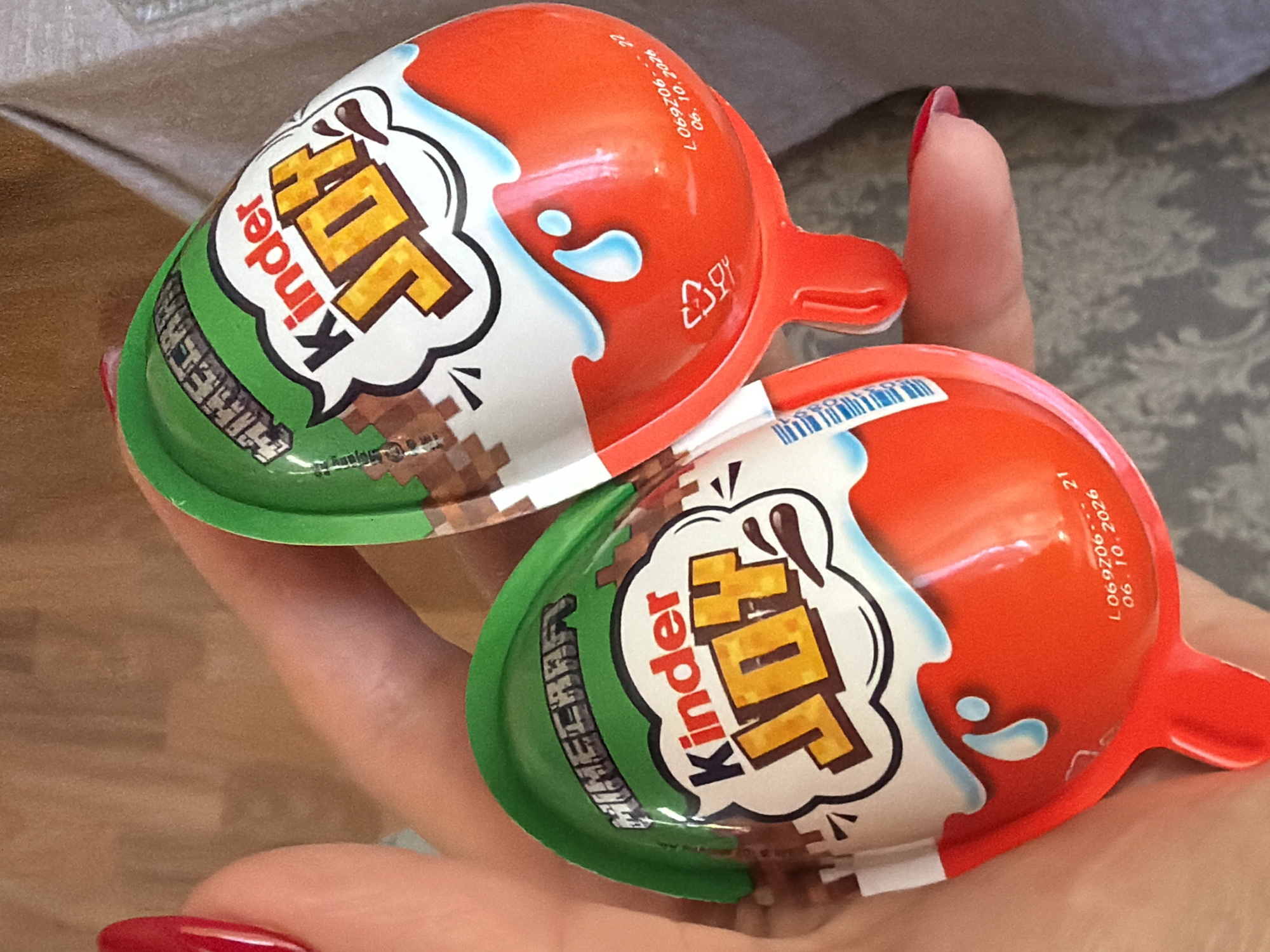Two Kinder Joy chocolate treats are displayed in a person's hand. The treats are Minecraft themed, with "Minecraft" imprinted on the green part of the packaging. The red tops show a white chocolate "splash". The packaging also shows the "Kinder Joy" logo. A production date of "06.10.2026" is visible on the red plastic top of the Kinder Joy package. The person holding the candies has manicured nails with red polish. The background shows a wooden surface and a patterned gray and white fabric. The location is not identifiable as Kamianske, Ukraine.