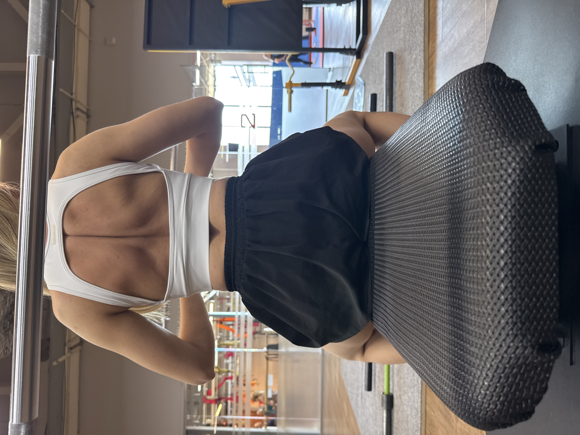A woman in athletic attire, a white sports bra and black shorts, is positioned at a gym. She appears to be using a machine. The gym setting features various equipment and visible workout spaces. The image was taken in Chaikovsky, Russia.