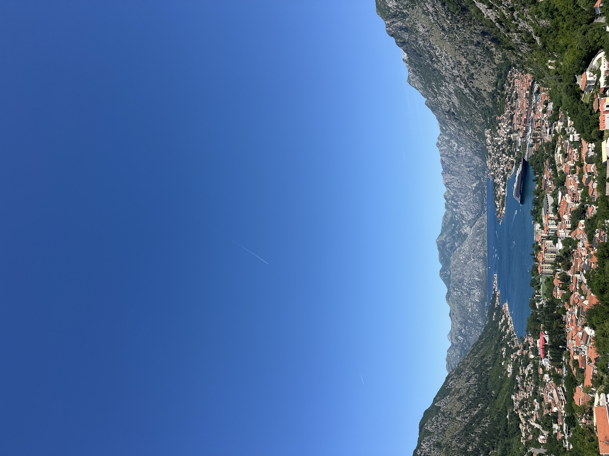 The image depicts a view of the Bay of Kotor in Škaljari, Montenegro. A clear blue sky occupies the majority of the frame. Prominent mountains frame the right side of the image, sloping down to meet the sea. The town of Škaljari sits along the coast, featuring terracotta-roofed buildings intermingled with greenery. A large cruise ship is visible in the bay. Two contrails are visible in the sky.