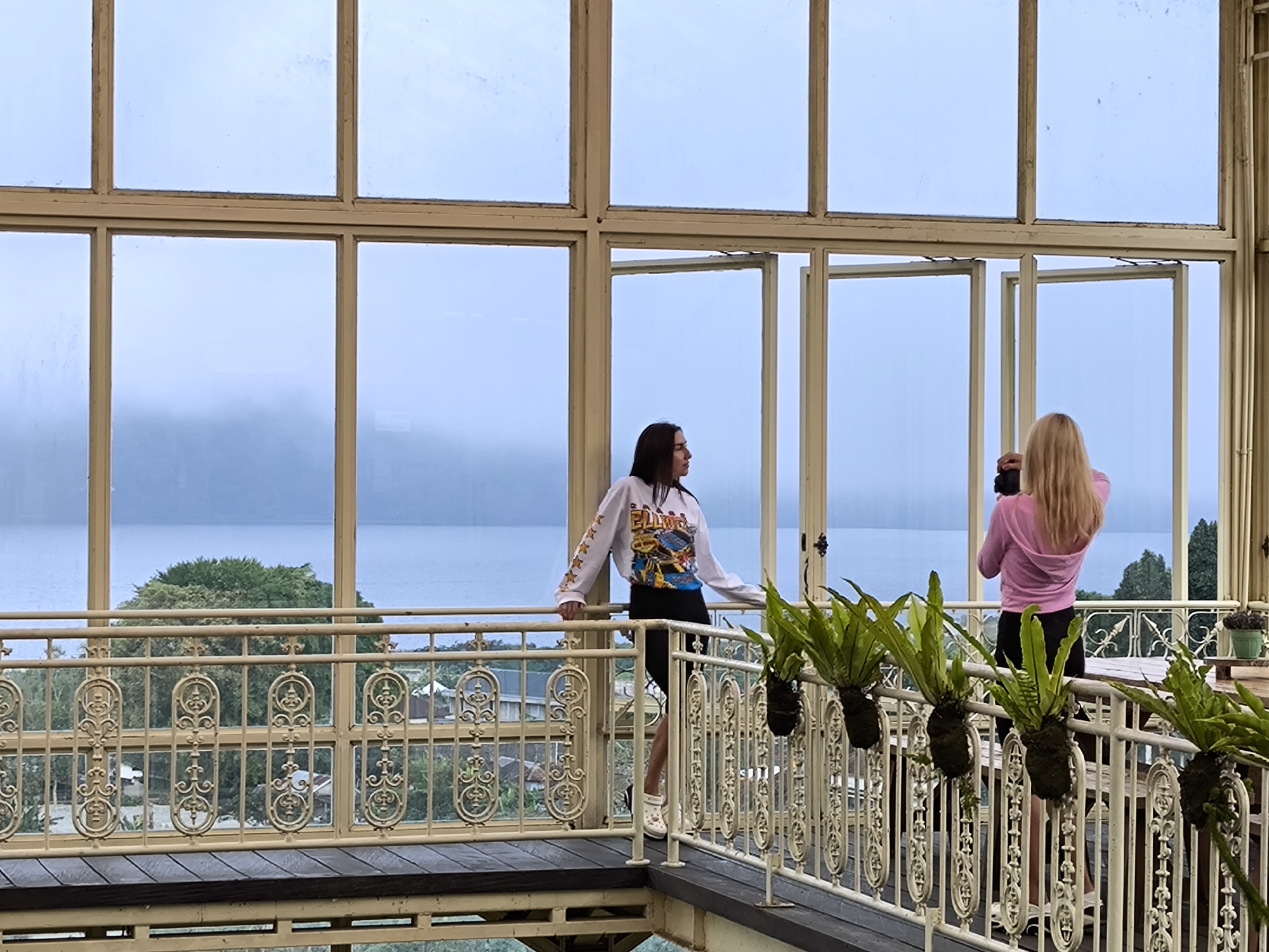 The image shows two women standing on a balcony, overlooking a lake. The women are wearing casual clothing. One woman has long dark hair, the other has long blonde hair and is holding a camera. The balcony has an ornate railing with hanging plants. The background features a lake with trees and a mountain in the distance. The scene is in Candikuning, Indonesia, as evidenced by the distinctive architectural style.