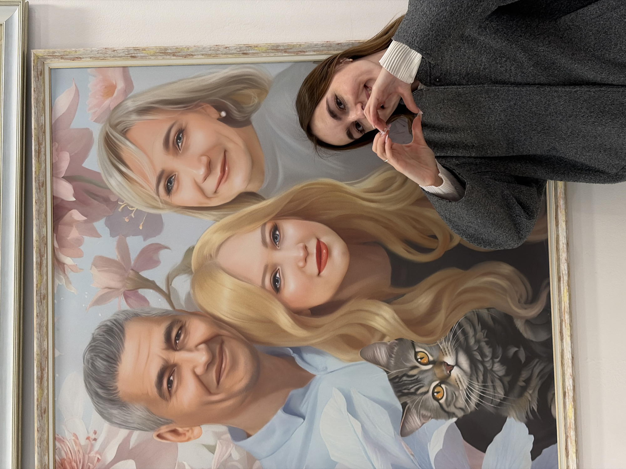 A framed portrait hangs on a wall, depicting a family of four with a cat. The father, with gray hair and a blue shirt, is at the bottom left. Above him is a blonde woman with red lipstick. To the left of her is an older blonde woman with a pearl earring. A gray tabby cat peeks out from behind the blonde woman. A young woman with brown hair and a gray jacket stands in front of the portrait, making a heart shape with her hands around a small, round object. The portrait is set in a decorative frame with gold accents. The wall is off-white. It is set in Izhevsk, Russia.