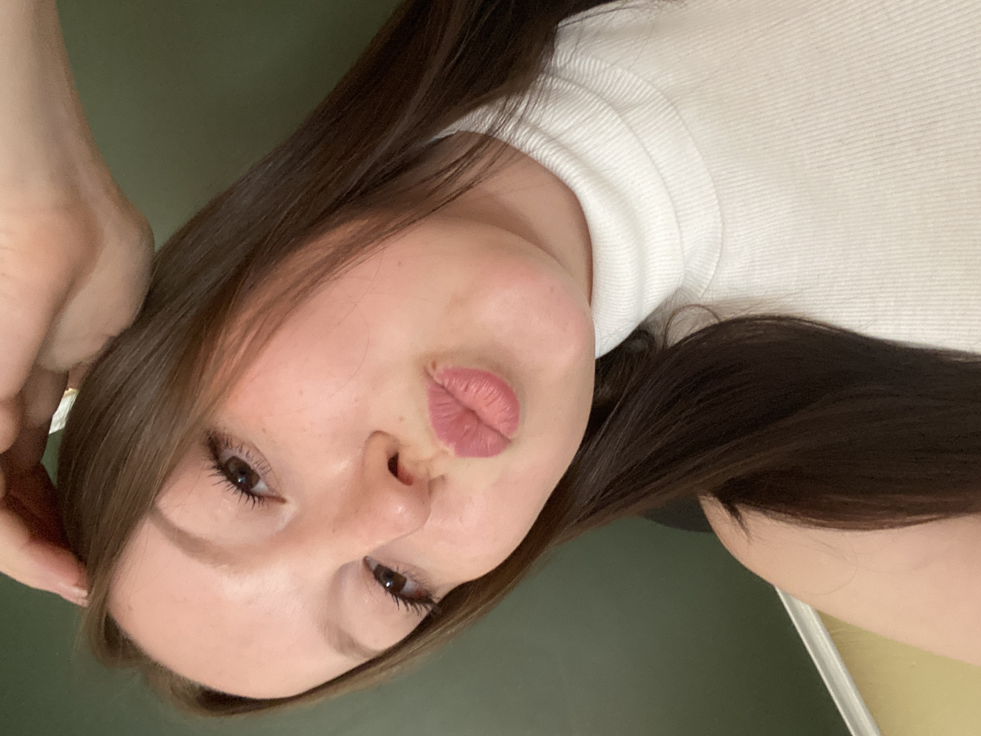 A woman with brown hair is posing for a selfie. She is wearing a white top and puckering her lips. The background is a dark green. The location is unknown, and there is no indication it is in Chaikovsky, Russia.