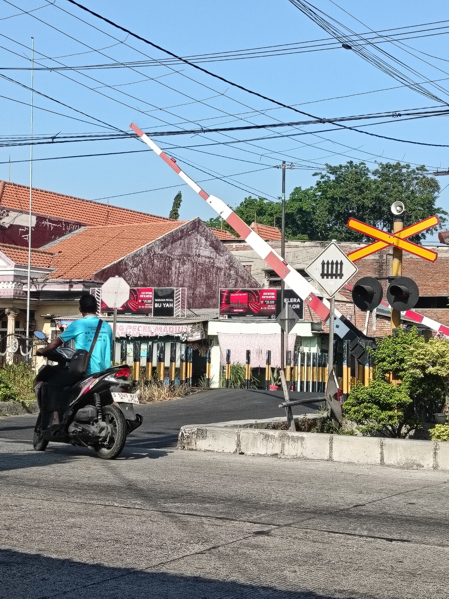 The image shows a street scene in Gedangan, Indonesia, with a focus on a railway crossing. In the foreground, a person wearing a blue t-shirt is riding a black scooter away from the camera, heading towards the railway tracks. The scooter has license plate number S 5745 NAF. The railway crossing features a striped barrier arm, which is partially lowered, and a yellow crossbuck sign indicating a railway crossing. There are also traffic signals and smaller warning signs. In the background, there are buildings with red tile roofs, a large tree, and numerous power lines crisscrossing the sky. The sky is clear and blue, suggesting a sunny day. The overall environment appears to be a typical urban or semi-urban area in Indonesia.