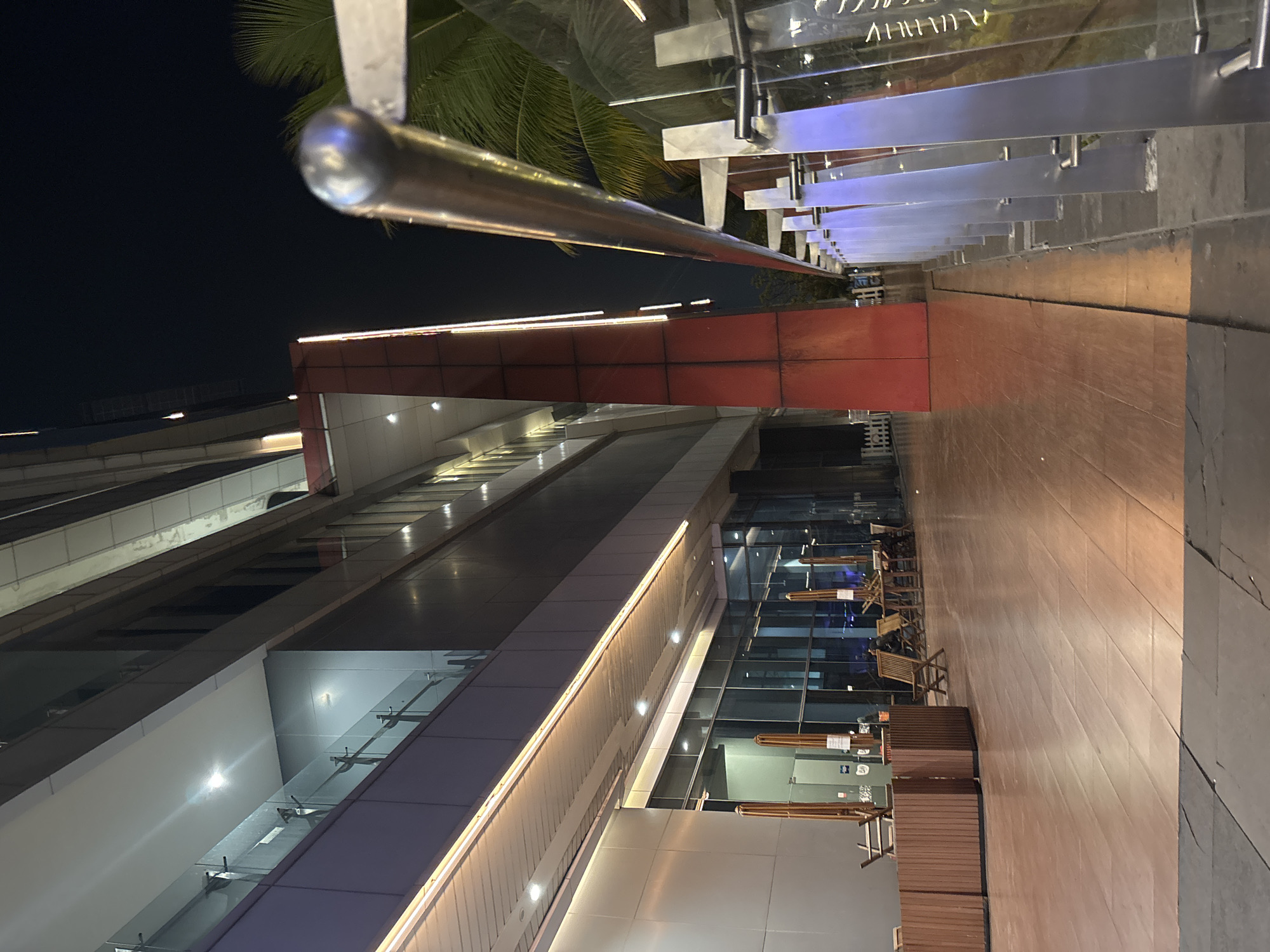 The image shows an outdoor seating area and a building at night. In the foreground, a polished metal railing runs along the right side of the frame, with a row of modern, illuminated metal posts supporting glass panels. The ground is paved with dark material that transitions to a reddish-brown wooden-like surface. To the left, several wooden tables and chairs are visible, some with umbrellas. The building behind the seating area has large glass windows and a modern facade with illuminated strips of light. A prominent red, angular architectural feature stands out against the building's darker exterior. Palm fronds are visible to the right, suggesting a tropical location. This scene is located at PIK Avenue Mall in North Jakarta, Indonesia. There are no people clearly visible in the image, and no specific actions or interactions are depicted.