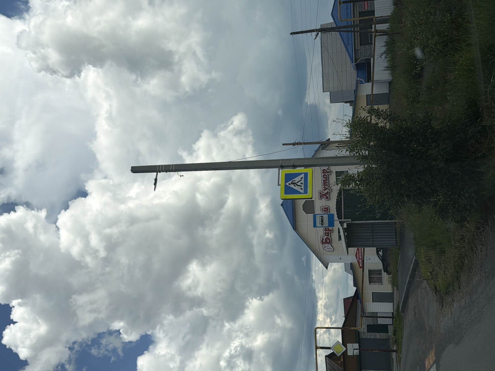 This image captures a street view in the city of Mezhevoy, Russia, under a dramatic, cloudy sky. In the foreground, a paved road curves to the left, with a grassy verge on the right. A pedestrian crossing is visible on the road.
Dominating the mid-ground is a two-story building with a blue roof, featuring signage that appears to read "Бари Хум" (Bari Hum) and another sign indicating "ПРОДУКТЫ МОЯ МЯСНО" (Products My Meat). Adjacent to this building is a dark green corrugated metal garage door.
To the left of the main building, a smaller, light-colored building with a yellow roof and a dark garage door is visible. Further left, a yellow square sign with a diamond shape is mounted on a yellow pipe.
A prominent concrete utility pole stands near the center of the image, supporting electrical wires and a street lamp. On this pole, a yellow pedestrian crossing sign is mounted. Another utility pole with wires is visible further to the right.
In the background, additional buildings with blue roofs and grey walls are present, along with a white fence. Dense green foliage and tall grass line the right side of the scene, obscuring parts of the buildings and the ground. A dark-colored car is parked near the entrance of one of the buildings. The overall atmosphere is overcast, with dark clouds suggesting an impending storm.