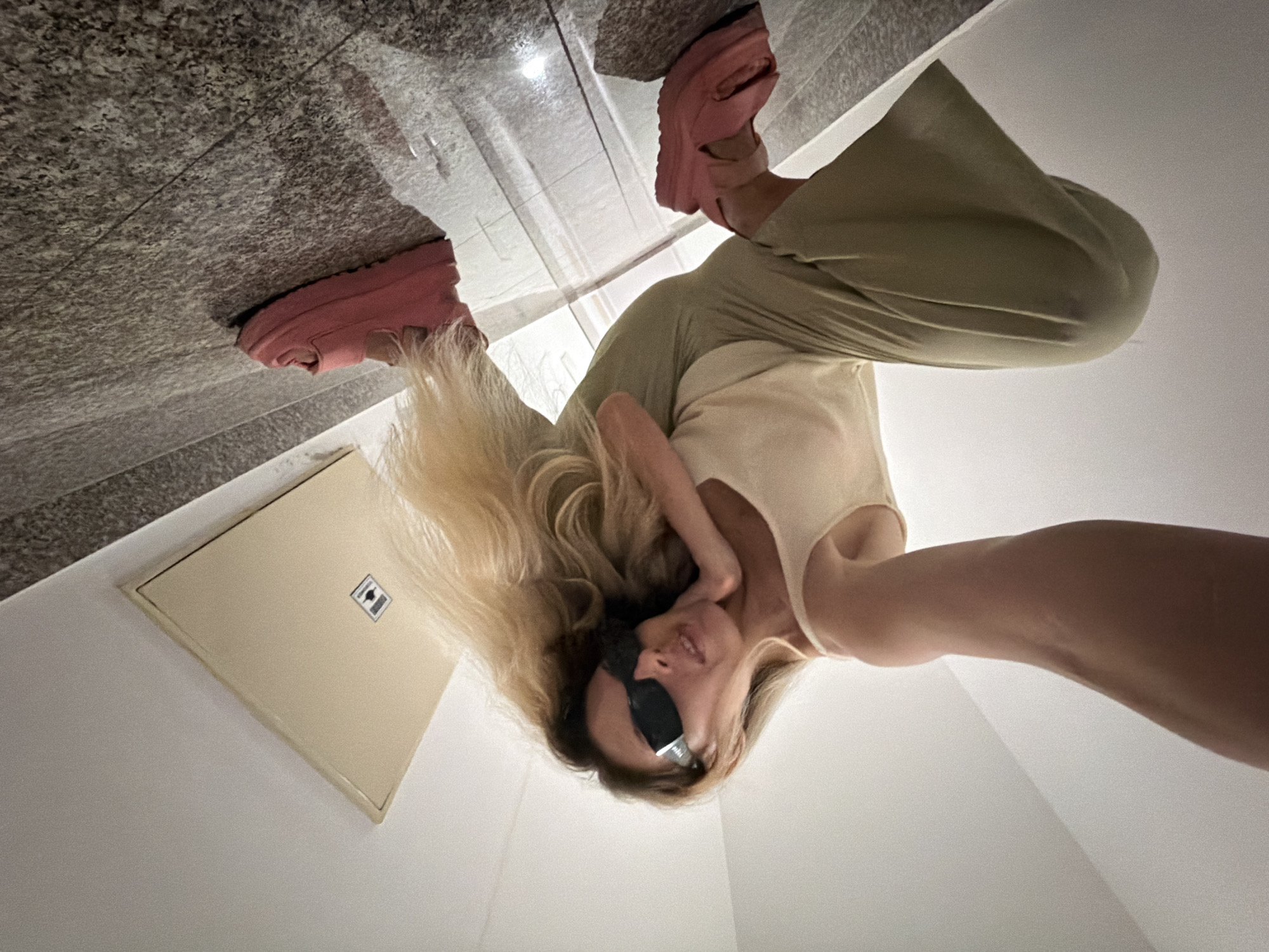 A woman with blonde hair and sunglasses is squatting on a stone floor in what appears to be a stairwell or hallway. She is wearing a light-colored tank top and loose-fitting green pants. She is also wearing pink platform sandals. A beige access panel is visible on the ceiling in the background. The lighting suggests an indoor environment, possibly in a residential building or hotel. The image is taken from a low angle, looking up at the woman.