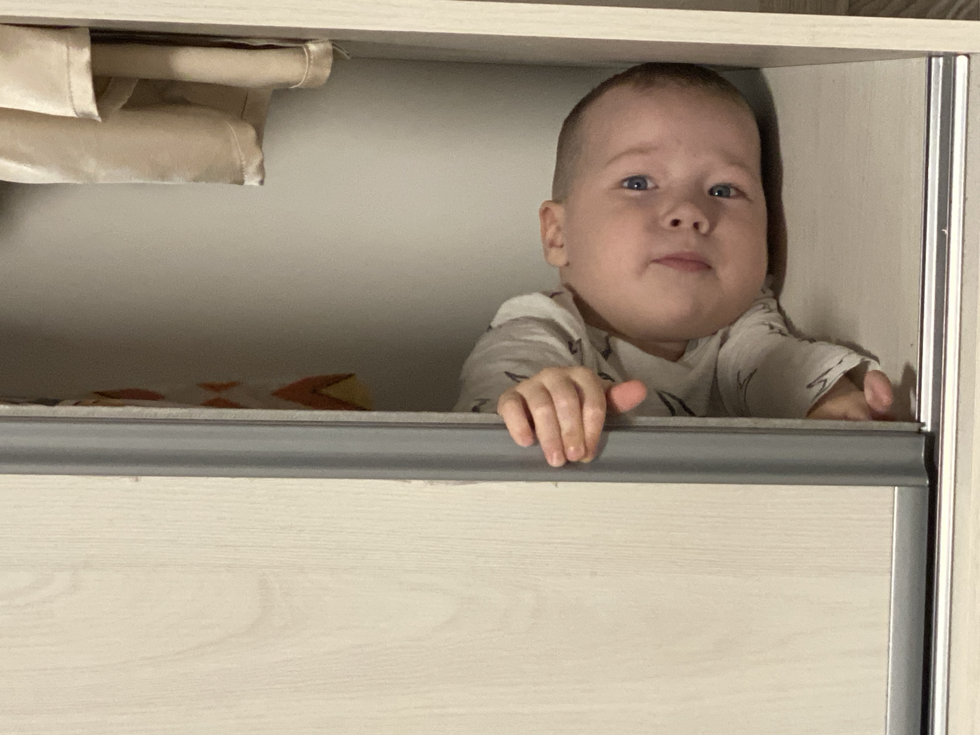 A baby is peering over the top of a drawer in a light-colored wooden cabinet. The baby has fair skin, blue eyes, and short, light brown hair. The baby is wearing a light-colored shirt with a pattern. The drawer is closed, and only the baby's head and hands are visible. There are some folded clothes or linens in the cabinet above the baby. The location is not identifiable as Sochi, Russia, from this image.