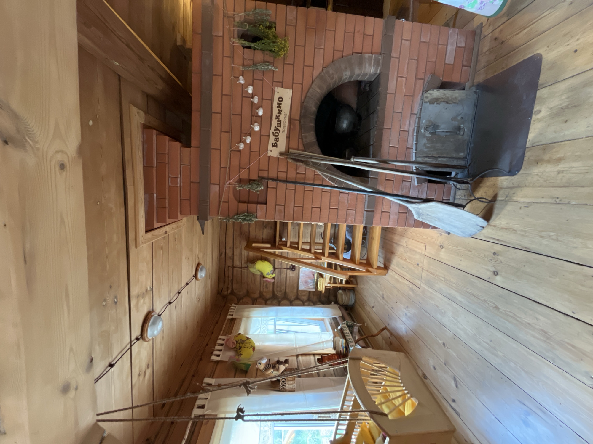 In a rustic, wooden interior in Vybolovo, Russia, a traditional Russian stove, or "pech," dominates the scene. Constructed of red brick, the stove features an arched opening containing a dark, cast-iron pot. Above the opening, a wooden sign reads "Бабушкино Подворье" (Grandmother's Courtyard), adorned with strings of dried herbs and garlic. To the left of the stove, a wooden scoop and a shovel rest on a metal ash pan. A wooden staircase leads upwards, partially obscured by hanging decorations. To the left of the stairs, a window with white curtains overlooks an outdoor scene. Near the window, a wooden cradle with yellow bedding is suspended by ropes. The room is furnished with wooden floors and log walls, and several decorative items, including stuffed toys and dried bunches of herbs, are visible. Lighting fixtures are mounted on the wooden ceiling.