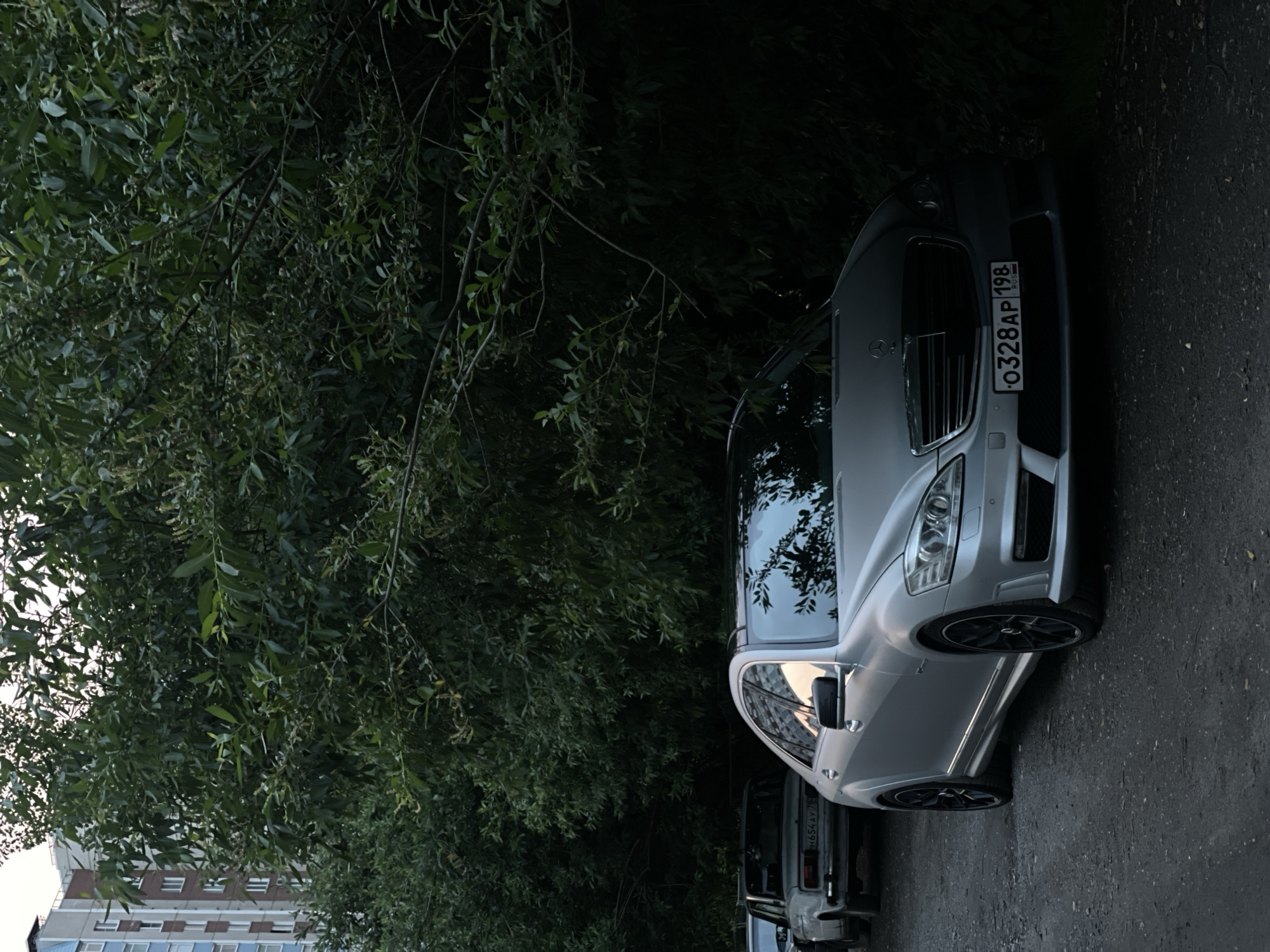 A silver Mercedes-Benz S-Class, with a black roof and black rims, is parked on a dirt road, partially obscured by dense green foliage. The car's license plate reads "O328AP 198 RUS". Behind the Mercedes, another older, lighter-colored car is visible. In the background, a multi-story apartment building with a light blue and brown facade stands behind a wall of trees. The scene appears to be an urban setting, possibly a residential area, with a mix of nature and man-made structures.