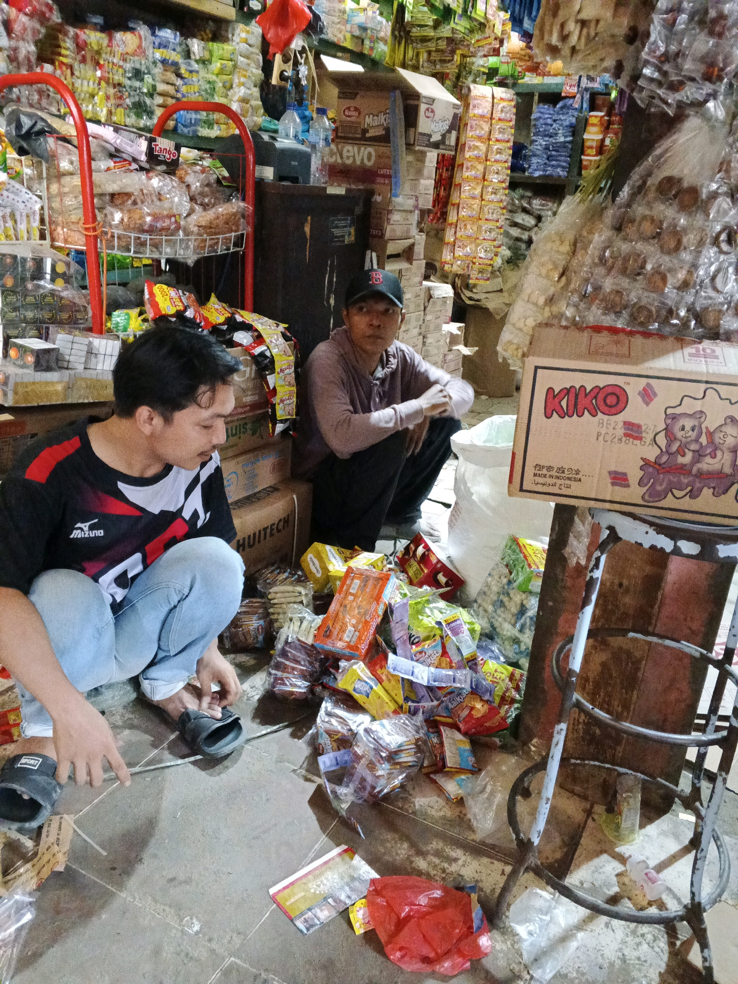 In Warungkiara, Indonesia, a market scene unfolds with two men amidst a display of goods. One man, wearing a black and red t-shirt with a white zigzag pattern and light blue jeans, is crouched down, examining something on the ground. He wears black slippers with "SPORT" visible on the left one. The second man, with a black baseball cap and a light purple long-sleeved shirt, is seated behind a pile of merchandise.
The environment is a bustling marketplace, evident from the stacked boxes, shelves laden with packaged food items, and various products arranged on the floor. The floor is a rough, textured concrete surface. Prominently displayed are boxes of "KIKO" cookies featuring cartoon bears, and a variety of bagged snacks and other consumables. A red metal cart is visible on the left, filled with more packaged goods. A white sack and a metal chair with a distressed surface add to the assortment of objects. The lighting suggests an indoor or covered market area, with shadows indicating diffuse light sources. The overall impression is one of commercial activity and the daily operations of a local market.