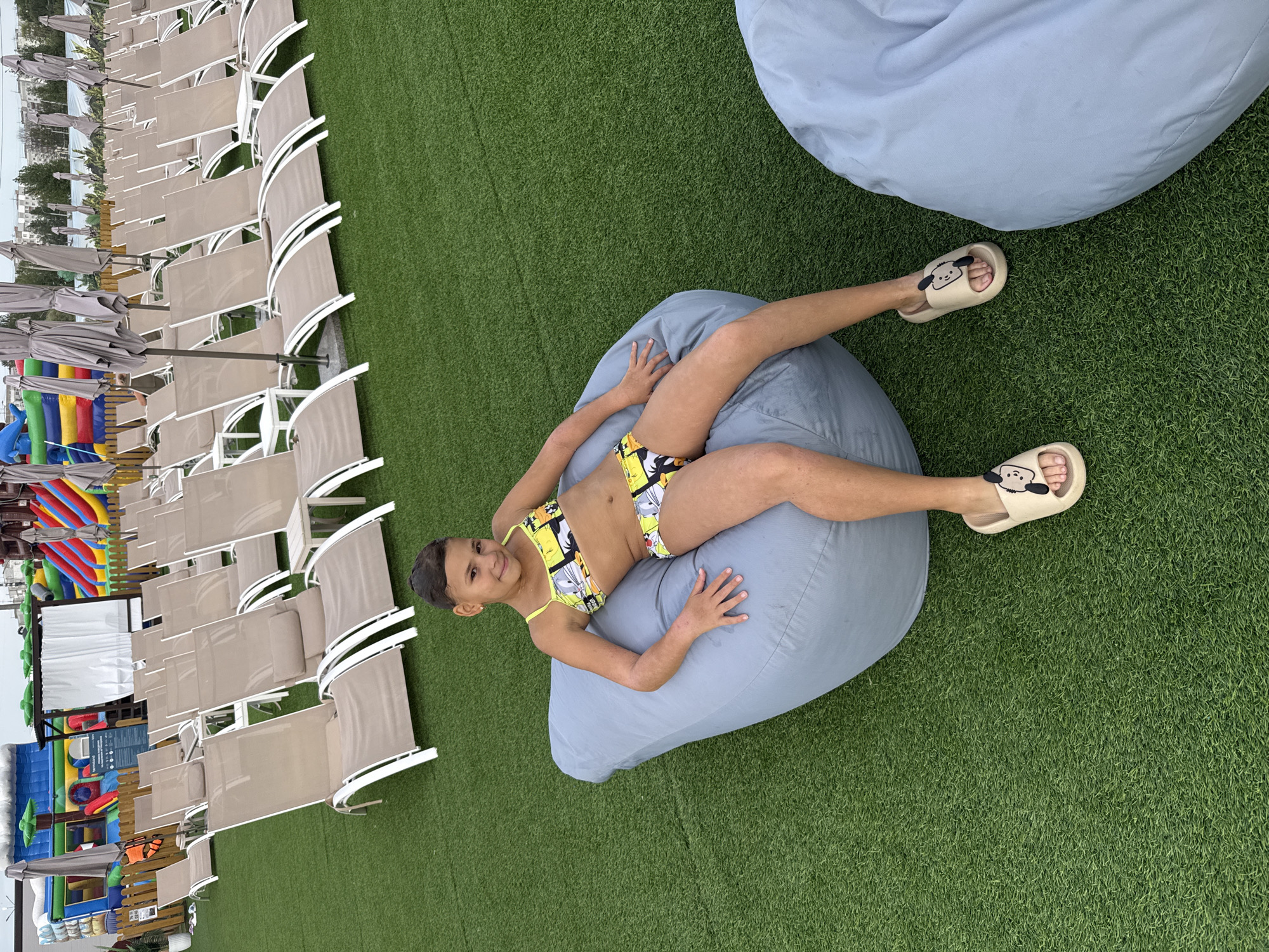 A young girl in a yellow bikini with a cartoon print lies on a large, light blue beanbag chair on a green artificial grass surface. She is on her back, with her legs extended and bent at the knees, and her arms resting on the beanbag. She is wearing light-colored slides with a cartoon face. The girl is smiling and looking towards the camera. In the background, rows of empty beige lounge chairs with umbrellas are lined up on the grass. To the left of the lounge chairs, there are colorful inflatable play structures. The scene appears to be at a resort or recreational area, likely during the daytime given the lighting. The overall mood is relaxed and cheerful. No visible text is present in the image.