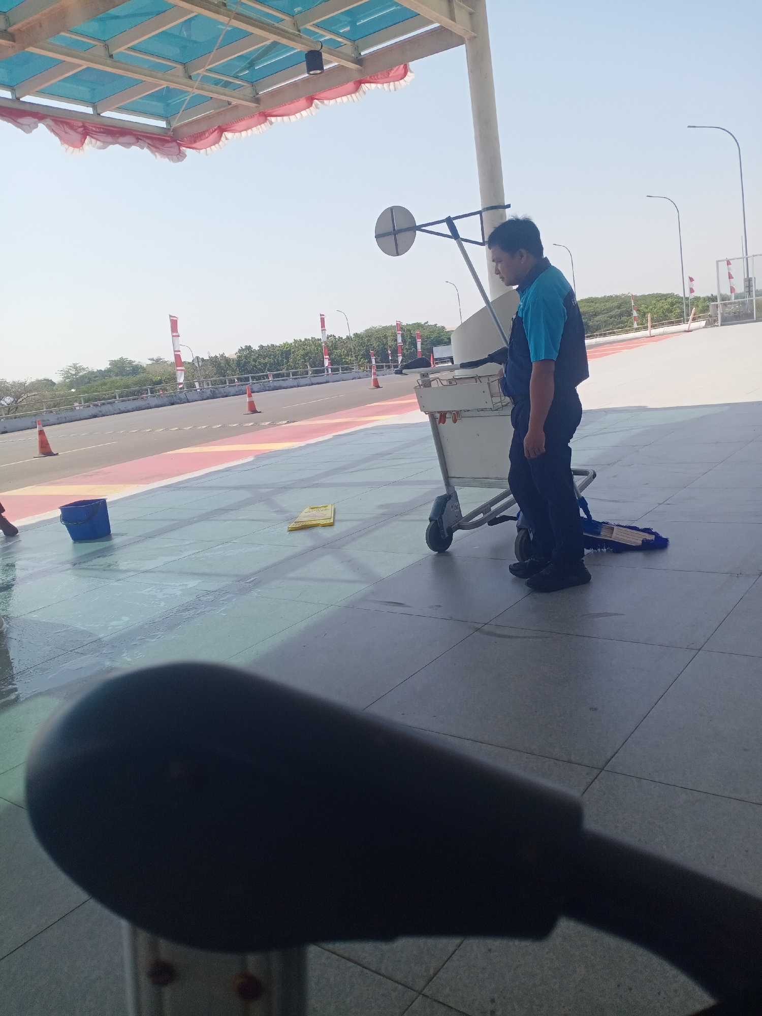 The image captures a scene outside what appears to be an airport terminal or a similar public transit hub in Baji Mangngai, Indonesia. The environment is characterized by a bright, clear sky and an open-air structure with a blue-paneled roof and red fabric trim. A man, dressed in a blue polo shirt and dark pants, stands next to a cleaning cart that holds a blue bucket and a mop. The ground is tiled and wet, suggesting recent cleaning activity. In the background, a road with orange traffic cones and several red and white flags is visible, along with trees and buildings. The overall impression is of an outdoor maintenance or cleaning operation in a public area.