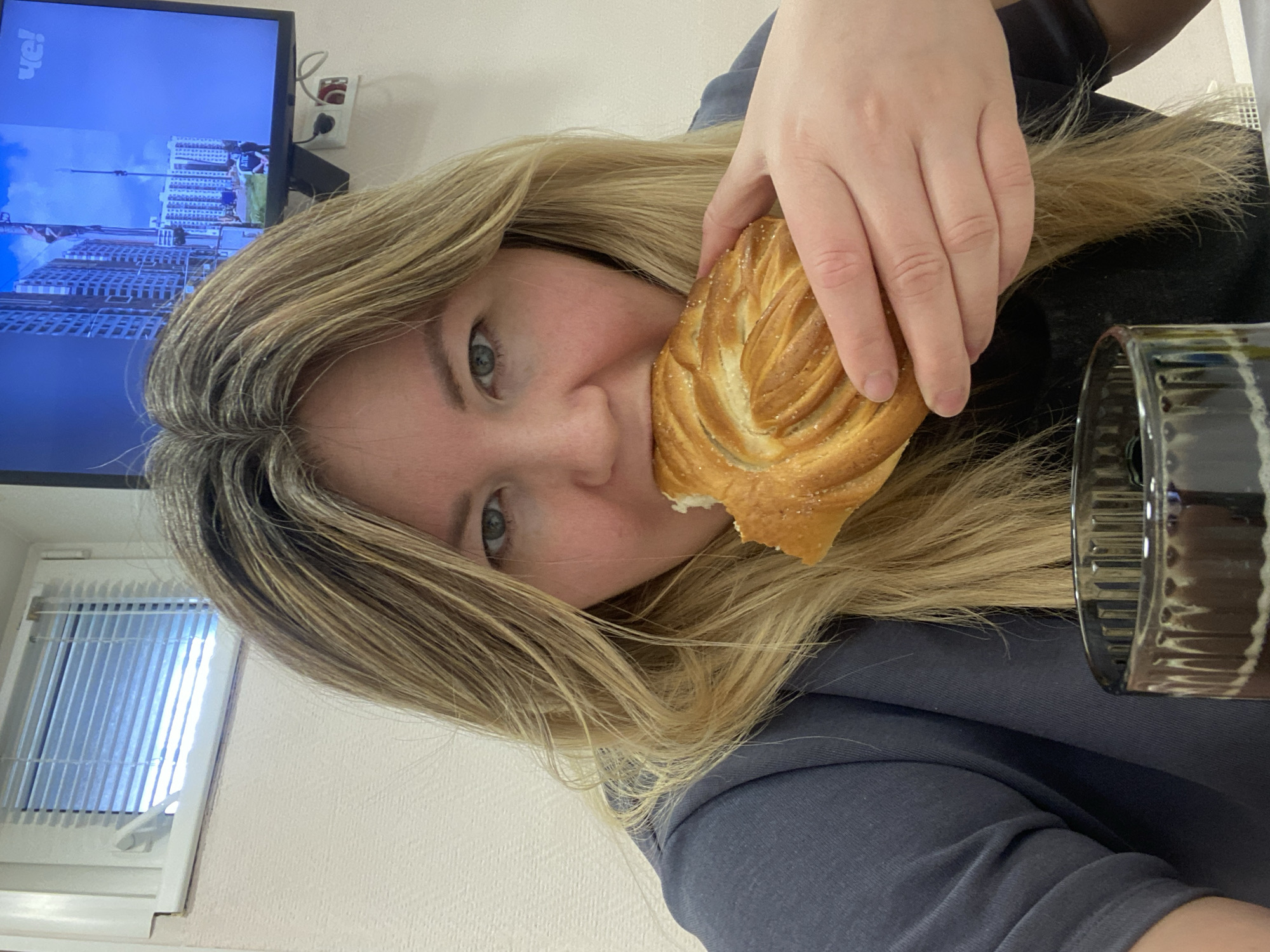 The image presents a close-up, rotated view of a woman, presumably indoors in Chaikovsky, Russia. The primary subject is a blonde woman, seen from a slightly low and tilted angle, with her left eye and part of her face visible. She is in the process of eating, holding a golden-brown, braided pastry or bun with her right hand and taking a bite from it. The pastry appears to have a glazed or sugary topping.
To her right, a dark, transparent glass or mug with a ribbed or fluted design is partially visible. The background reveals details of the room: a flat-screen television is mounted on a wall on the left, displaying an image of modern city buildings under a blue sky. Prominently visible on the television screen is the text "ЧЕ!", indicating the Russian TV channel "Che". Next to the TV, an electrical outlet with a red plug is attached to the wall. In the lower left corner of the image, a window with white vertical blinds is partially seen. The walls of the room are a light, plain color. The woman's expression appears neutral, capturing a casual moment of eating. The overall perspective of the image is tilted, suggesting it might be a selfie or a photo taken at an unusual angle.