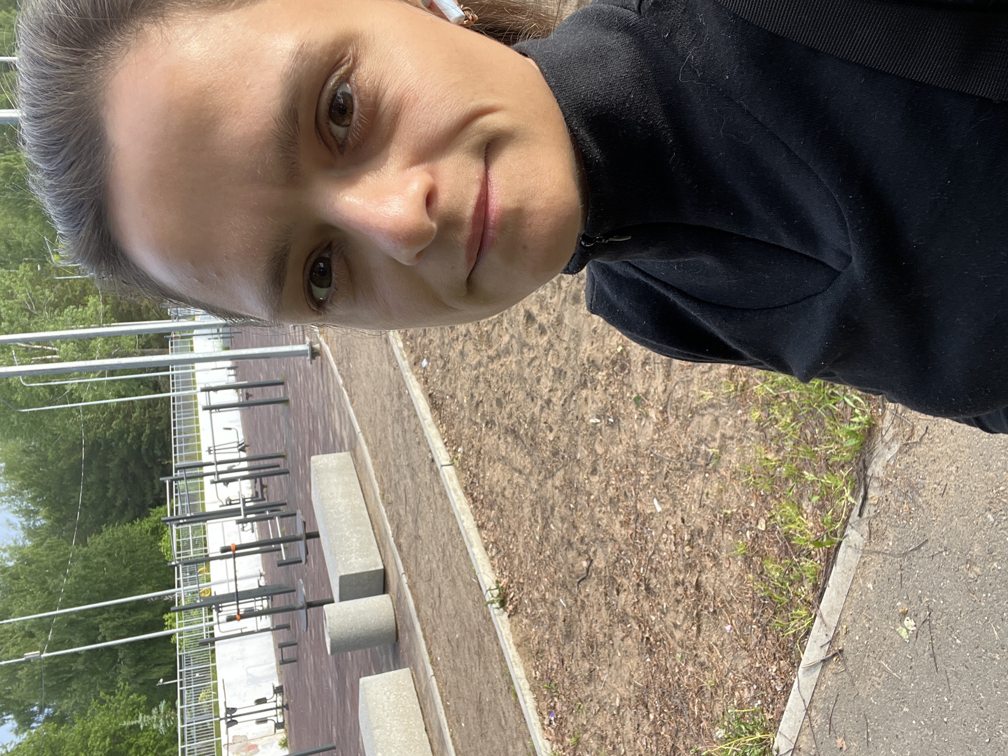 A selfie of a woman with fair skin and brown hair pulled back, wearing a black sweatshirt. The background shows an outdoor workout area in the city of Chaikovsky, Russia. The area includes metal exercise equipment and concrete benches on a paved surface. A dirt area with sparse vegetation borders the paved area. Trees and a fence are visible in the distance.