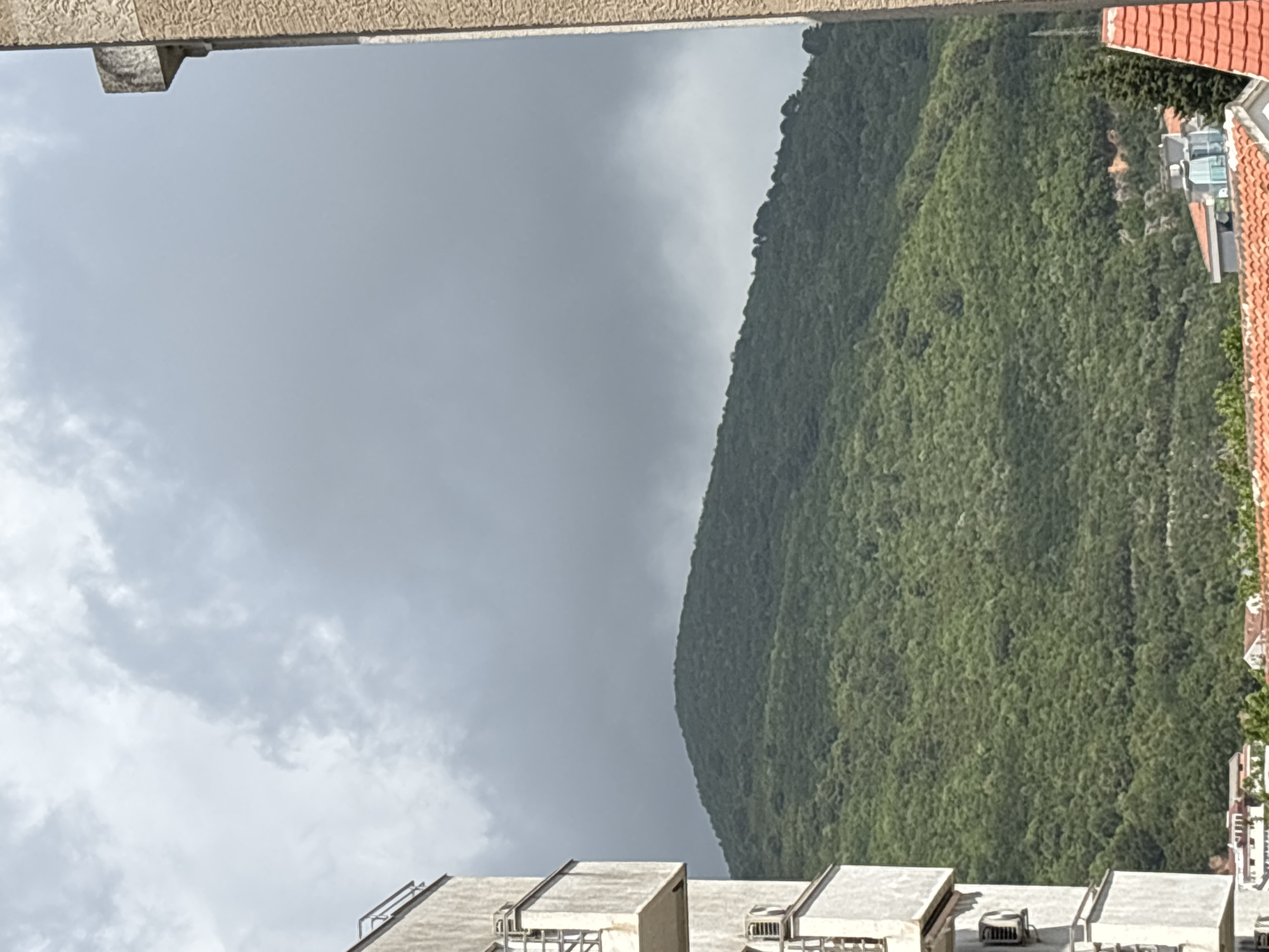 Overcast view of Budva, Montenegro. A green mountain rises to the right. Gray clouds fill the left sky. Buildings with orange tile roofs are nestled near the mountain base. Modern apartment buildings with rooftop features are visible at the bottom of the frame. A concrete ledge is at the top edge.