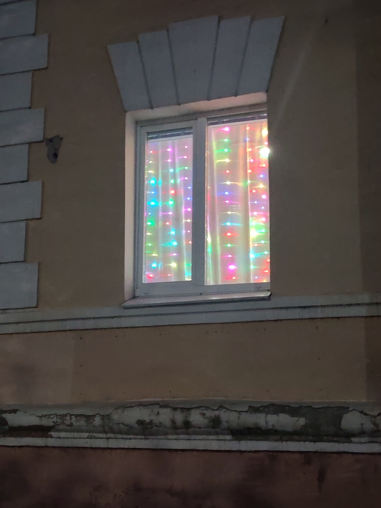 The image shows a close-up view of a window on a building's exterior. The window is divided into two panes, and behind the glass, a string of colorful LED lights is draped across a white curtain. The lights are a mix of pink, blue, green, and yellow, creating a festive and illuminated effect. The exterior wall of the building is a muted tan color, with decorative stone-like elements above and below the window. The bottom portion of the image shows a dark, textured surface, possibly a wall or foundation, with some peeling paint or plaster. The scene appears to be captured at night, with the bright lights of the window being the primary source of illumination. There is no indication of the specific location within the city of Vladimir, Russia, or the presence of any people.