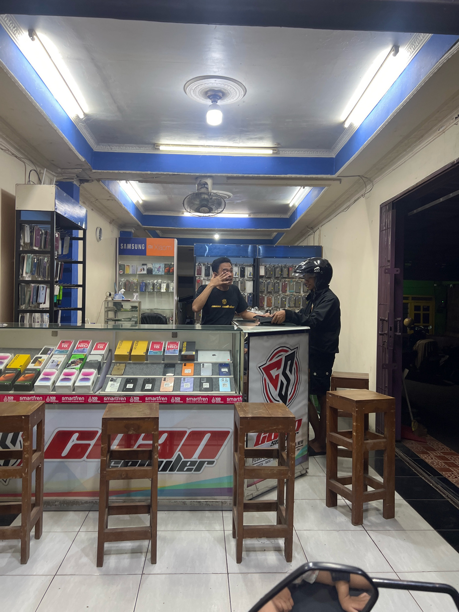 In Kecamatan Pati, Indonesia, an interior scene depicts a small electronics shop at night. The shop is illuminated by fluorescent lights on the ceiling, with blue accents along the ceiling border. Two men are present. One man, wearing a black t-shirt with a yellow logo and dark pants, stands behind a counter and appears to be speaking, gesturing with his right hand near his face. The other man, wearing a black jacket, black pants, and a black motorcycle helmet, stands on the other side of the counter, facing the first man and holding an object that resembles a laptop or tablet.
The shop features a glass display counter filled with boxed mobile phones and accessories. Behind the counter, shelves are stocked with more phone cases and accessories. A prominent display on the wall shows "SAMSUNG" and "XIAOMI" logos above rows of phone accessories. Two wooden stools are placed in front of the counter. The floor is tiled with white squares. The shop's entrance, a dark doorway, is visible to the right, with a glimpse of an outdoor area and a motorcycle parked outside.