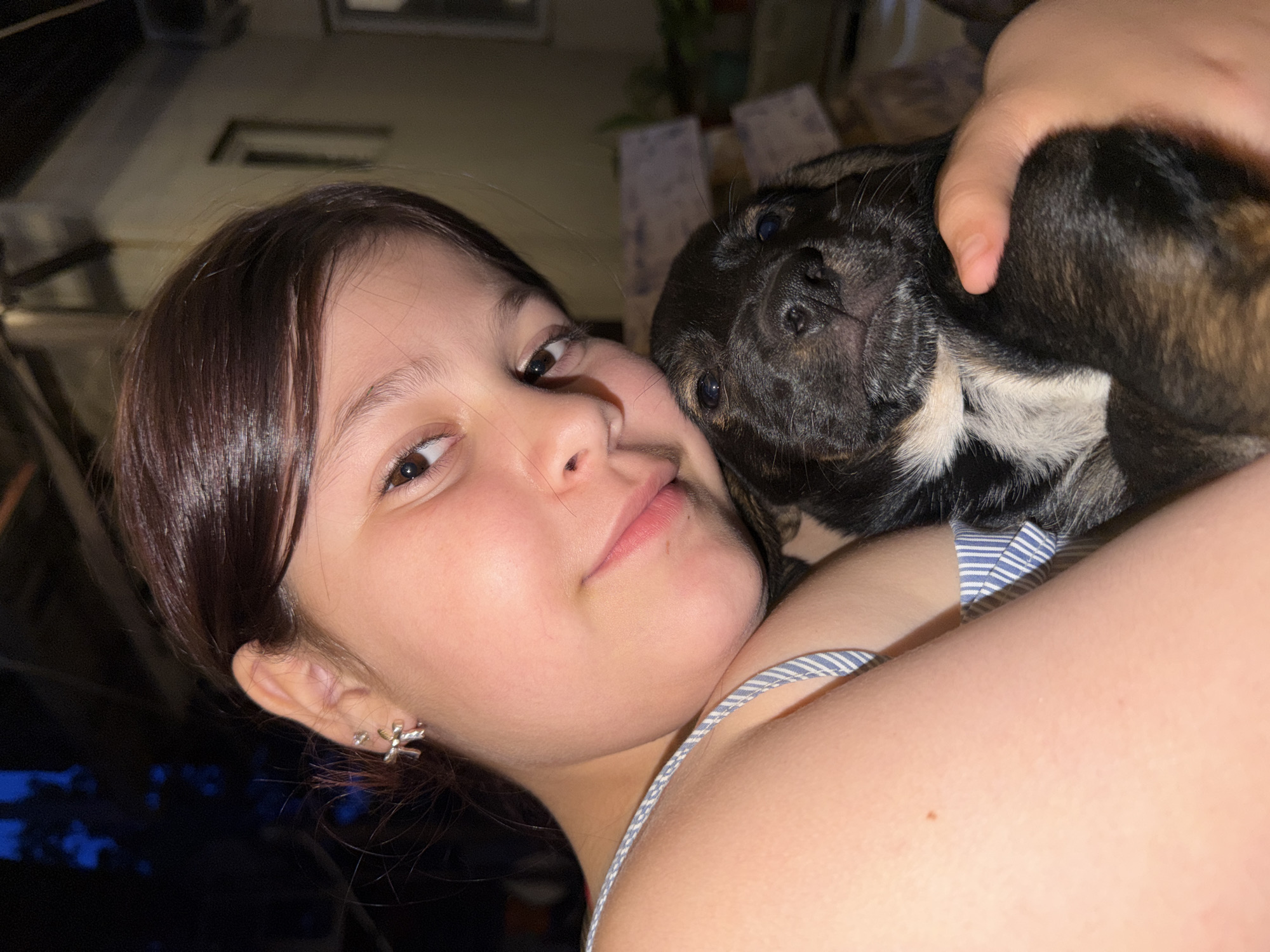 A young girl with dark hair and a pale complexion is holding a black and brown puppy. She is wearing a blue and white striped top and a silver earring in her left ear. The puppy's head is resting on her chest, and its body is partially visible. The background is dark and out of focus, suggesting an indoor setting at night, possibly in Tuapse, Russia. There are hints of wooden structures and some faint light sources in the background. The overall impression is one of tenderness and affection between the girl and the puppy.