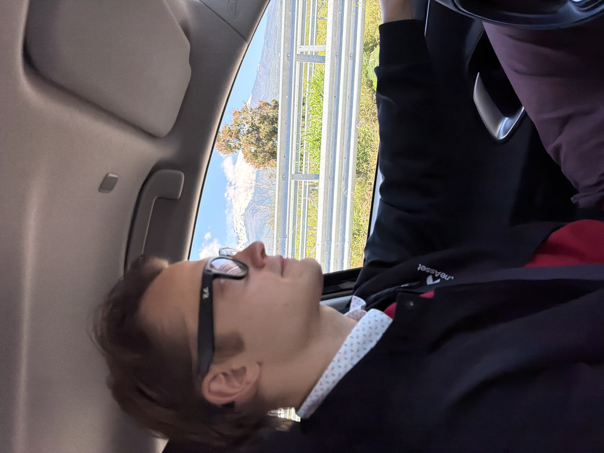 The image depicts a young adult male resting or sleeping as a passenger inside a car during daytime. He is positioned with his head against the car's headliner, looking towards the outside view through the window. He wears black-framed glasses, a dark jacket over a white collared shirt with a red pattern, and a red undershirt. Part of maroon-colored pants is also visible. The jacket features a white logo and the reversed text "neAsset" on the sleeve.
The car's interior shows a grey headliner, a sun visor, and a grab handle. Through the window, a bright outdoor landscape is visible, featuring a metallic multi-bar guardrail running horizontally. Beyond the guardrail, there is green vegetation and a tree. In the distance, large mountains, possibly with snow or clouds on their peaks, rise under a clear blue sky with scattered white clouds. The word "AIRBAG" is visible on the car's interior pillar next to the window. The scene suggests a journey through a scenic, mountainous region, aligning with the context of Santa Venerina, Italy. The bright lighting indicates it is daytime.