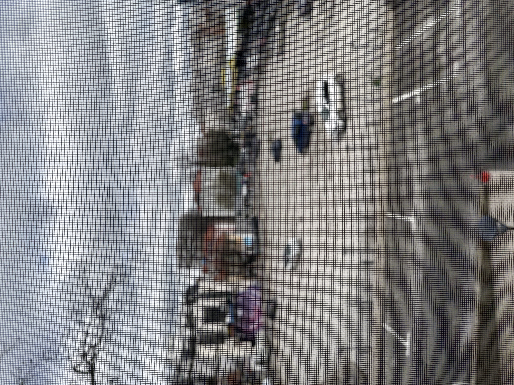 The image presents an outdoor urban scene in Pleven, Bulgaria, observed through a fine mesh screen, which imposes a grid pattern and contributes to the overall blurriness. The sky is overcast, dominated by shades of light gray and white, with bare tree branches visible against it in the upper left.
The mid-ground is occupied by a large, open paved area, consistent with a parking lot or public square. Several vehicles are parked on this surface. A light-colored sedan is positioned in the lower-mid right, with a dark blue or black sedan situated further back and to its upper left. Another light-colored vehicle is visible further into the mid-ground on the left. White painted lines mark parking spaces on the pavement, and several small, dark, vertical cylindrical objects, possibly bollards, are intermittently placed across the area.
In the background, a series of multi-story urban buildings line the far edge of the paved area. These structures are indistinct, appearing in muted tones of gray, brown, and off-white. One building in the left-mid background displays a large, blurry area of purplish-pink coloration, suggesting a mural or graffiti. Beyond these buildings, additional vehicles, some appearing yellow or light-colored, are discernible on what appears to be another street or parking zone.
The immediate foreground features a dark, indistinct horizontal band at the bottom, likely a windowsill or railing. A small, blurry red object and a darker triangular shape are partially visible on the bottom right edge of this foreground element. No discernible people or specific actions are present in the scene.