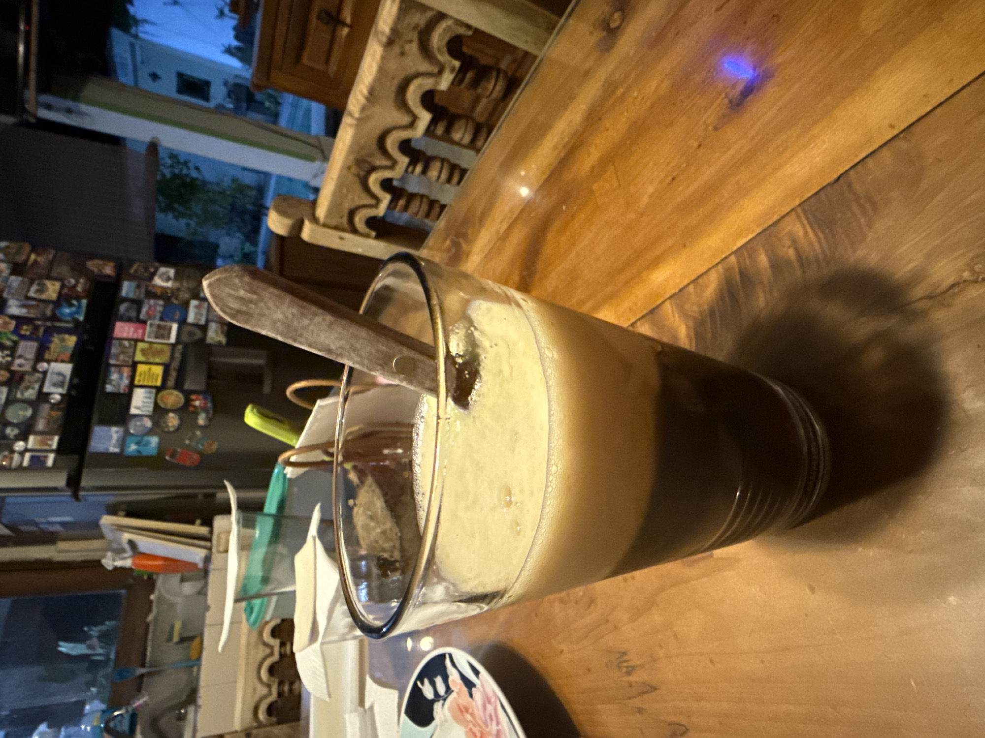 A glass of beverage sits on a wooden table. The drink appears to be a dark liquid at the bottom and a foamy, light-colored substance on top. A wooden spoon is inside the glass. Behind the glass are miscellaneous kitchen objects and supplies. A refrigerator with numerous magnets is visible in the background. The window behind the refrigerator offers a view of the outside. The wooden table upon which the drink sits is heavily detailed. A carved wooden chair back is nearby. The location in the city of Xochitepec, Mexico, is indeterminable.