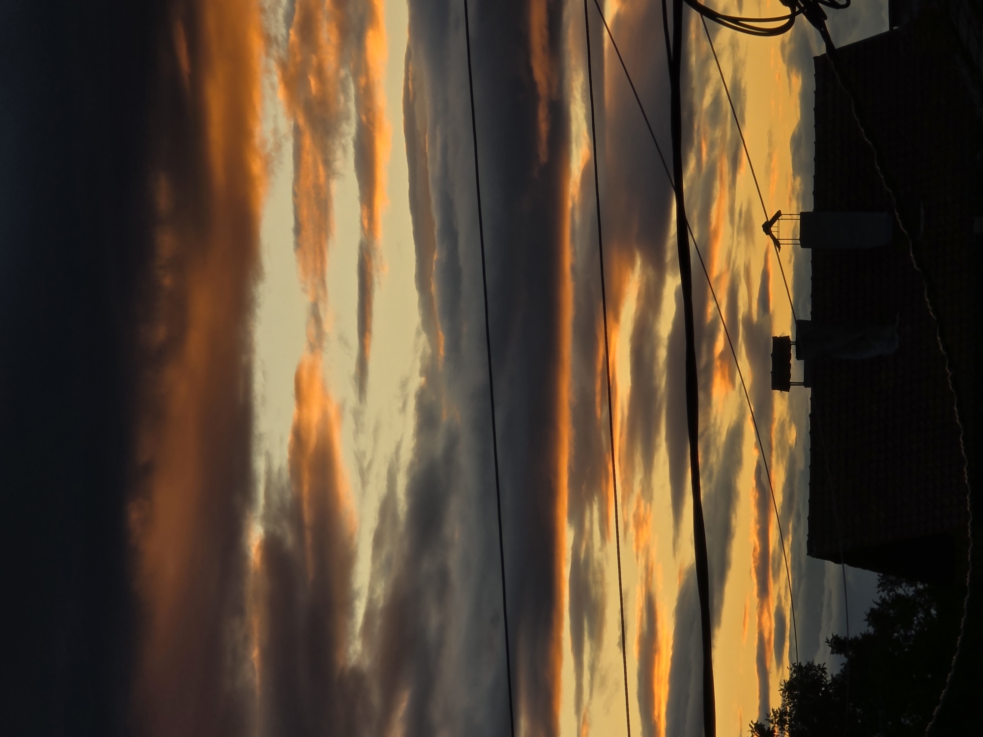 The image depicts a sunset in Yagodovo, Bulgaria. The sky is streaked with orange and gray clouds. Power lines and utility poles are visible in the foreground, silhouetted against the colorful sky. The outline of a building and some trees are visible in the lower right corner. The photo is taken from a low angle, looking up towards the sky. The image is vertically oriented.