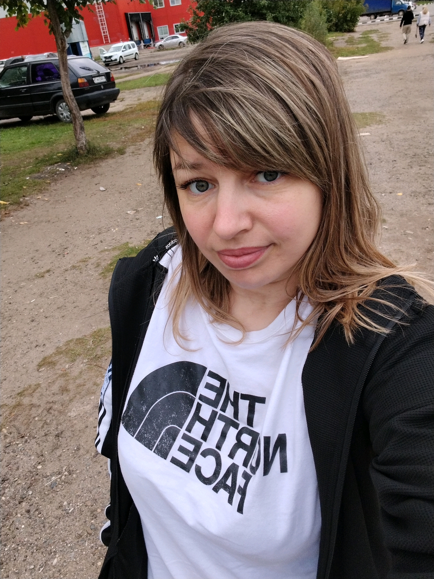 A woman with light brown hair and blue eyes is taking a selfie outdoors. She is wearing a white t-shirt with a black graphic print and a black jacket. In the background, there is a red building, a black car, and other vehicles. The ground is a dirt and gravel surface. The scene appears to be a parking lot or an industrial area, possibly in Yaroslavl, Russia.