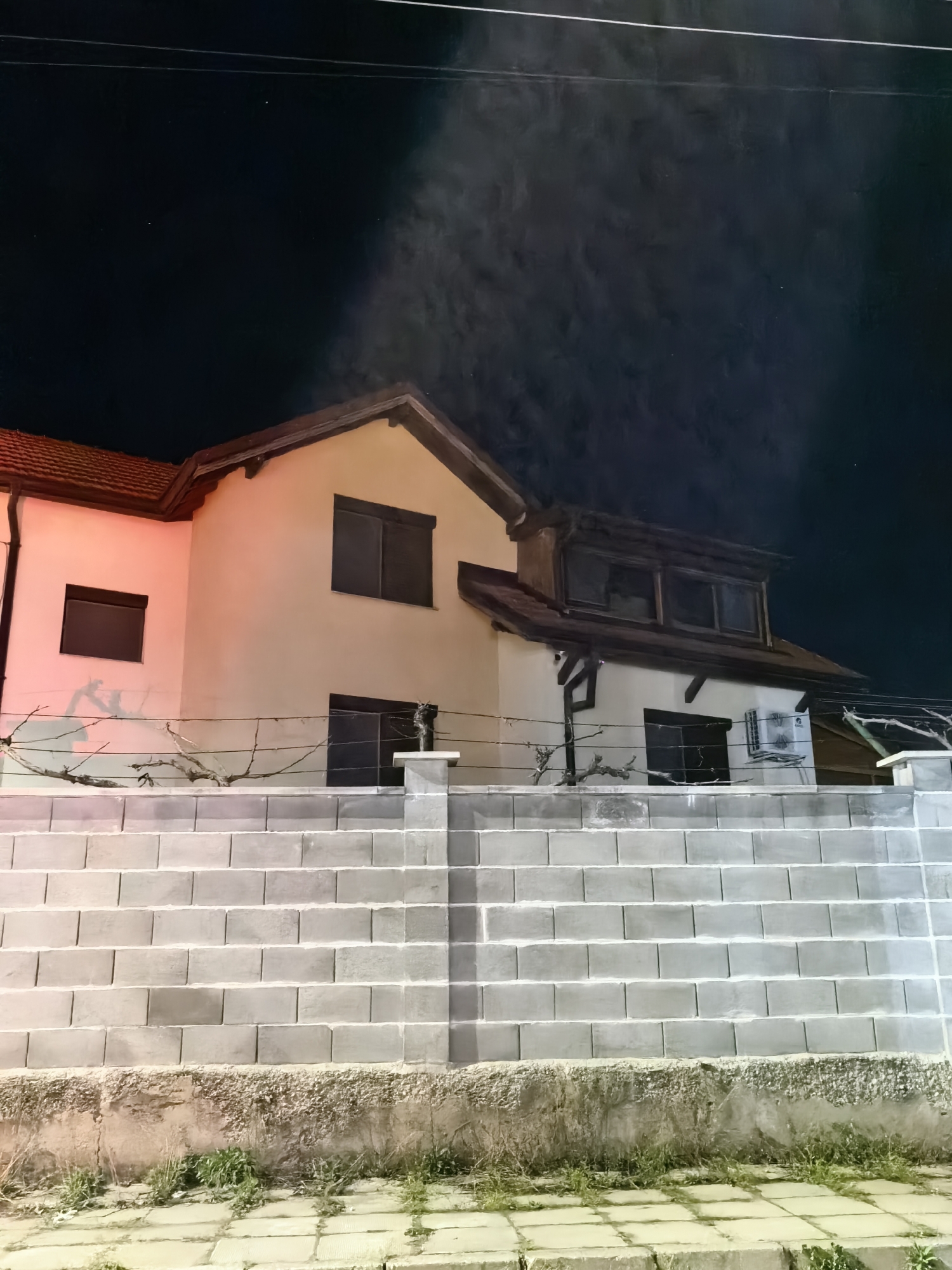 Captured at night in an unidentified city, the photograph showcases a two-story building behind a substantial, stepped concrete block wall. The building has a light-colored facade with contrasting dark window frames and a brown tiled roof. There is an additional structure with a gable window on the second story of the building's right side. A few thin, leafless branches are visible in front of the building, strung along a wire fence atop the wall. A light source highlights a section of the structure's facade and the wall, casting a faint glow. The sky above is dark, punctuated with what appear to be faint atmospheric conditions. The ground is paved with stone blocks, and small patches of grass are visible at the base of the wall.