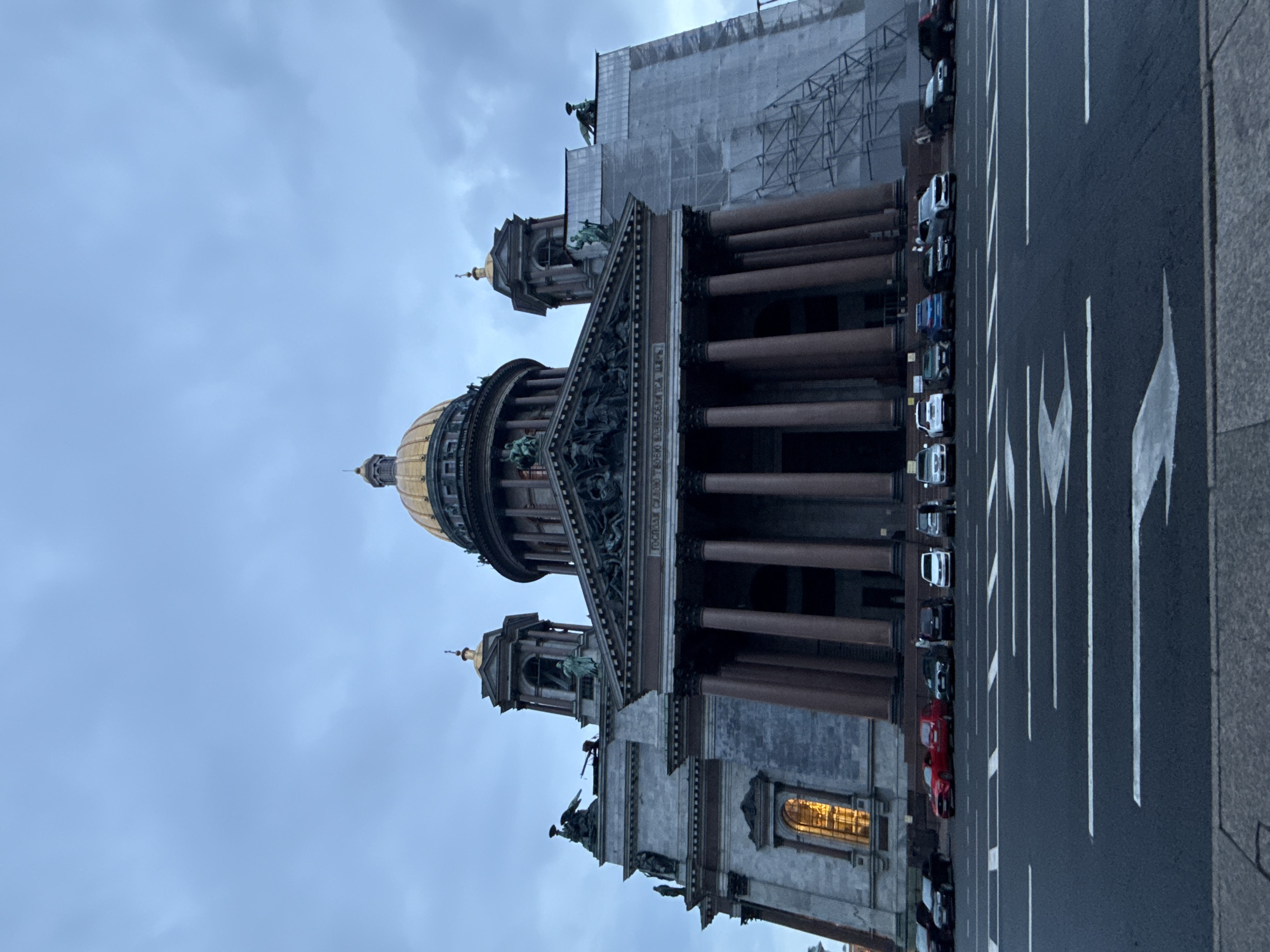 The image captures a low-angle view of a grand domed building, likely a cathedral, under a cloudy sky. The building features classical architecture with a large golden dome, prominent columns, and intricate sculptures and carvings adorning its facade. Statues of figures can be seen on the building's exterior and atop some of its towers. To the right of the main structure, scaffolding and protective sheeting cover a portion of the building, suggesting ongoing renovation or construction work. In the foreground, a wide road with clear lane markings is visible, with several cars parked along the side. The lighting suggests it is either early morning or late evening. No people are clearly distinguishable in the image.