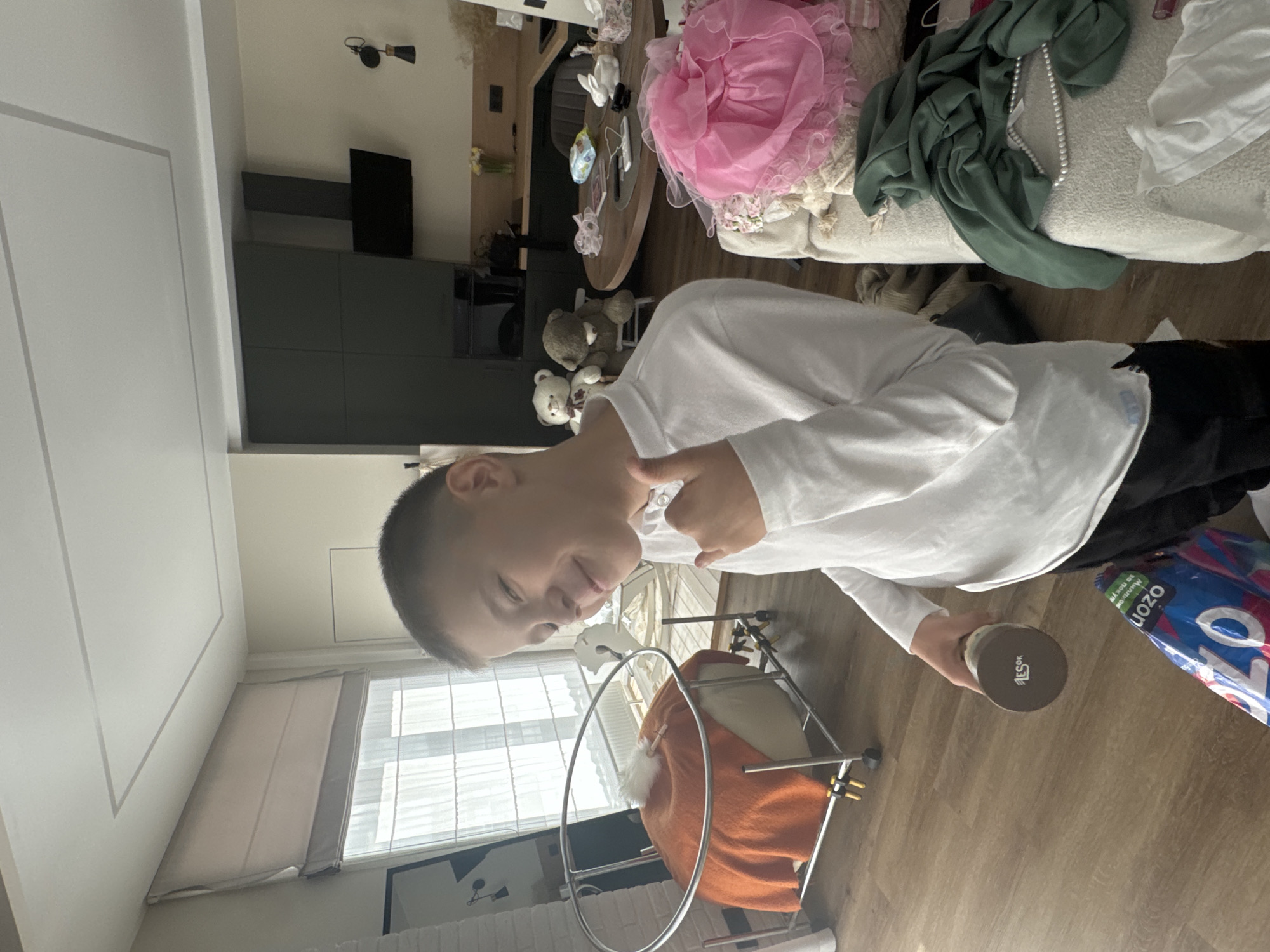 A boy in a white long-sleeved shirt and black pants is holding a dark-colored container with text on the bottom while standing on a wooden floor. He is pointing with his right index finger. Behind him is a baby swing with an orange blanket. A window with white blinds is visible in the background. To the left of the window, there's a white wall with a gray trim, and a glimpse of a television mounted on the wall. In the distance is a living room with a couch covered in clothes and toys, including a pink dress, a green garment with pearls, and several teddy bears on a small table. A dining table is also visible. The location is Izhevsk, Russia.