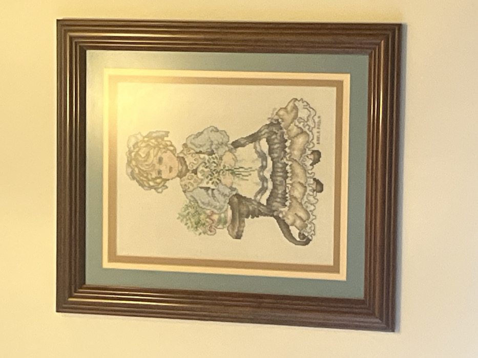 A dark brown, multi-ridged rectangular picture frame is mounted on a plain, light beige wall in Mexico City, Mexico. The frame contains an artwork double-matted with a wide light blue-green border and a thin inner cream border. The artwork is a color illustration depicting a young girl with voluminous curly blonde hair, fair skin, and youthful features. She wears a light blue long-sleeved dress, a white ruffled apron, and a dark, multi-tiered ruffled skirt. The girl stands centrally, holding a small bouquet of white flowers. To her left, a dark brown, circular-topped pedestal table supports a small potted plant with green foliage. A faint dark signature, "BARLA PROLA," is visible in the bottom right corner of the illustration. A prominent glare from a light source is present across the upper right quadrant of the framed image.
