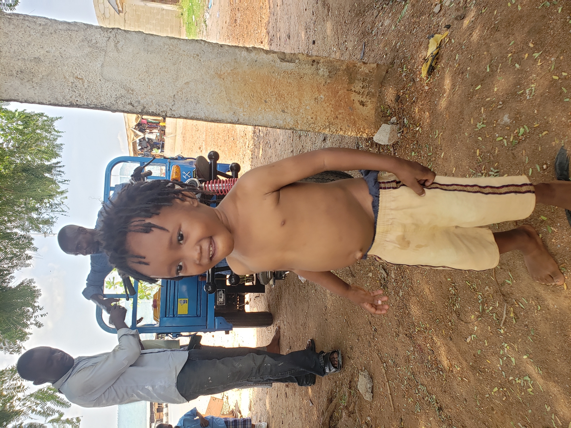 Adankolo, Nigeria. A shirtless child with dreadlocks stands in front of a blue tricycle with two adults. One adult wears a light grey shirt and dark jeans, while the other wears a blue shirt. The ground is dirt and sand. A concrete structure is in the background.
