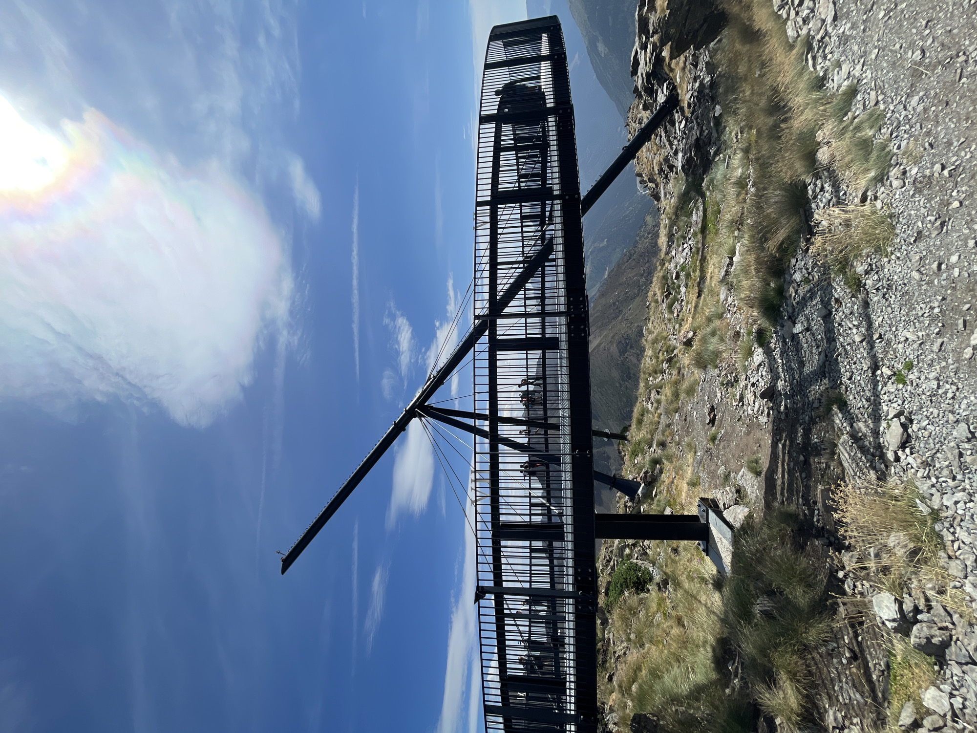 An outdoor, eye-level, medium shot captures a panoramic viewpoint extending from a mountain with a circular platform and railing system, overlooking a valley filled with hazy, distant mountains under a bright, partly cloudy sky. The viewpoint structure is made of black metal and is supported by a thick pole. The platform has a black railing and a walkway made of metal grates. Several people are visible on the platform. The terrain surrounding the viewpoint is rocky with sparse, dry grass. The sky is blue with scattered white clouds, and a rainbow-colored atmospheric phenomenon is visible in the upper right portion of the sky.