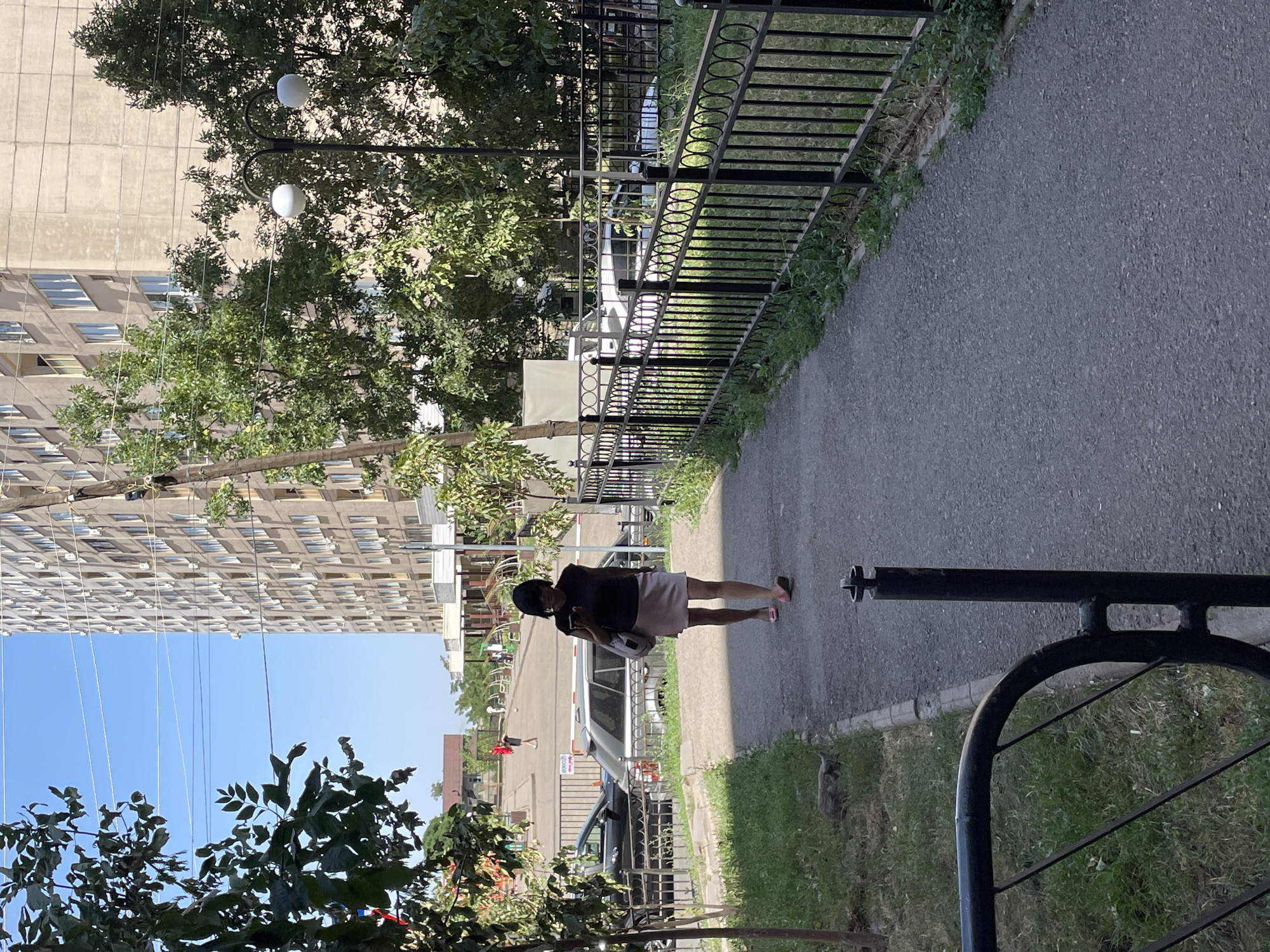 A woman with short black hair, wearing a dark sleeveless top and a light pink skirt, walks on a paved sidewalk. She is looking down at her phone, which she holds in her right hand. She has a light-colored shoulder bag slung over her left shoulder and wears pink open-toed sandals. She is walking away from the camera, heading towards a street lined with buildings and trees.
The scene appears to be a residential area in the city of Almaty, Kazakhstan. In the background, there is a tall, light-colored apartment building with numerous windows and air conditioning units. A black metal fence runs along the right side of the sidewalk, separating it from a grassy area with trees. Several parked cars are visible beyond the fence, including a silver SUV and a dark-colored sedan. In the distance, a few people are visible in an open courtyard area, one of whom is wearing a red shirt and appears to be kicking a ball. The sky is clear and blue. A stray cat is lying on the grass to the left of the sidewalk. A lamppost with two spherical lights is visible among the trees.