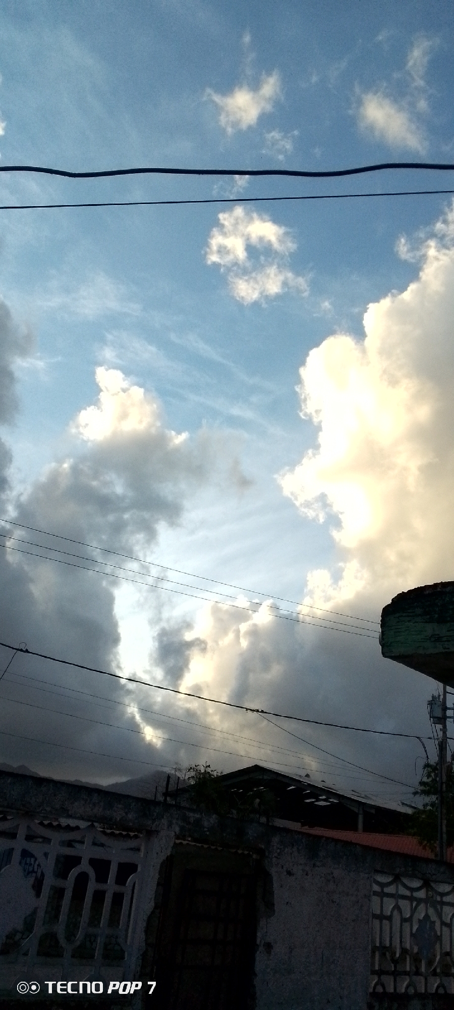 The image depicts a sky scene in Altagracia, Venezuela, with a mix of white and dark clouds against a blue sky. Several electrical wires traverse the upper portion of the frame. In the lower part of the image, a building with a patterned wall and a partially visible roof is present. A small portion of a green structure is visible on the right. The image appears to be taken during the day, though the lighting suggests it might be late afternoon or approaching evening due to the shadows and contrast in the clouds. A mountain is barely visible in the background, between the roof and the clouds. The logo of "TECNOP POP 7" is present in the bottom left corner.