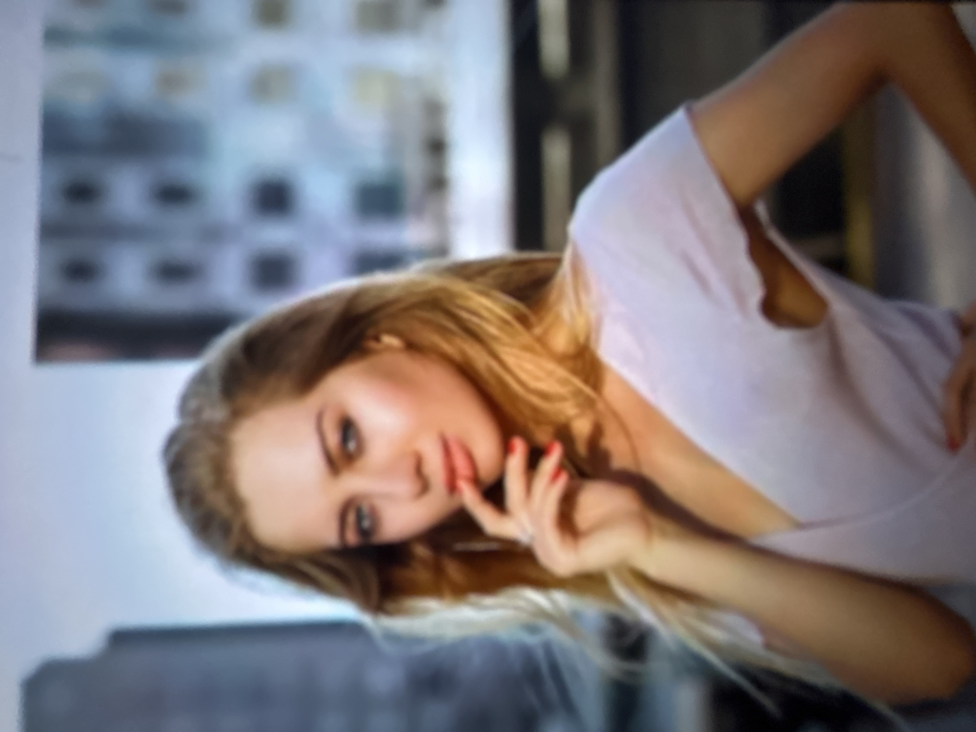 An outdoor, slightly blurred, medium-shot photograph features a young Caucasian woman with blonde hair, a fair complexion, and blue eyes. She is wearing a light purple, V-neck t-shirt. Her right hand is raised to her mouth, with her index finger touching her lips. Her fingernails are painted red. She is wearing a ring on her left ring finger. The background is out of focus, showing indistinct urban buildings and a bright sky. The woman is likely in a city, but the location of Gelendzhik, Russia, cannot be determined from the visual information provided in the image.