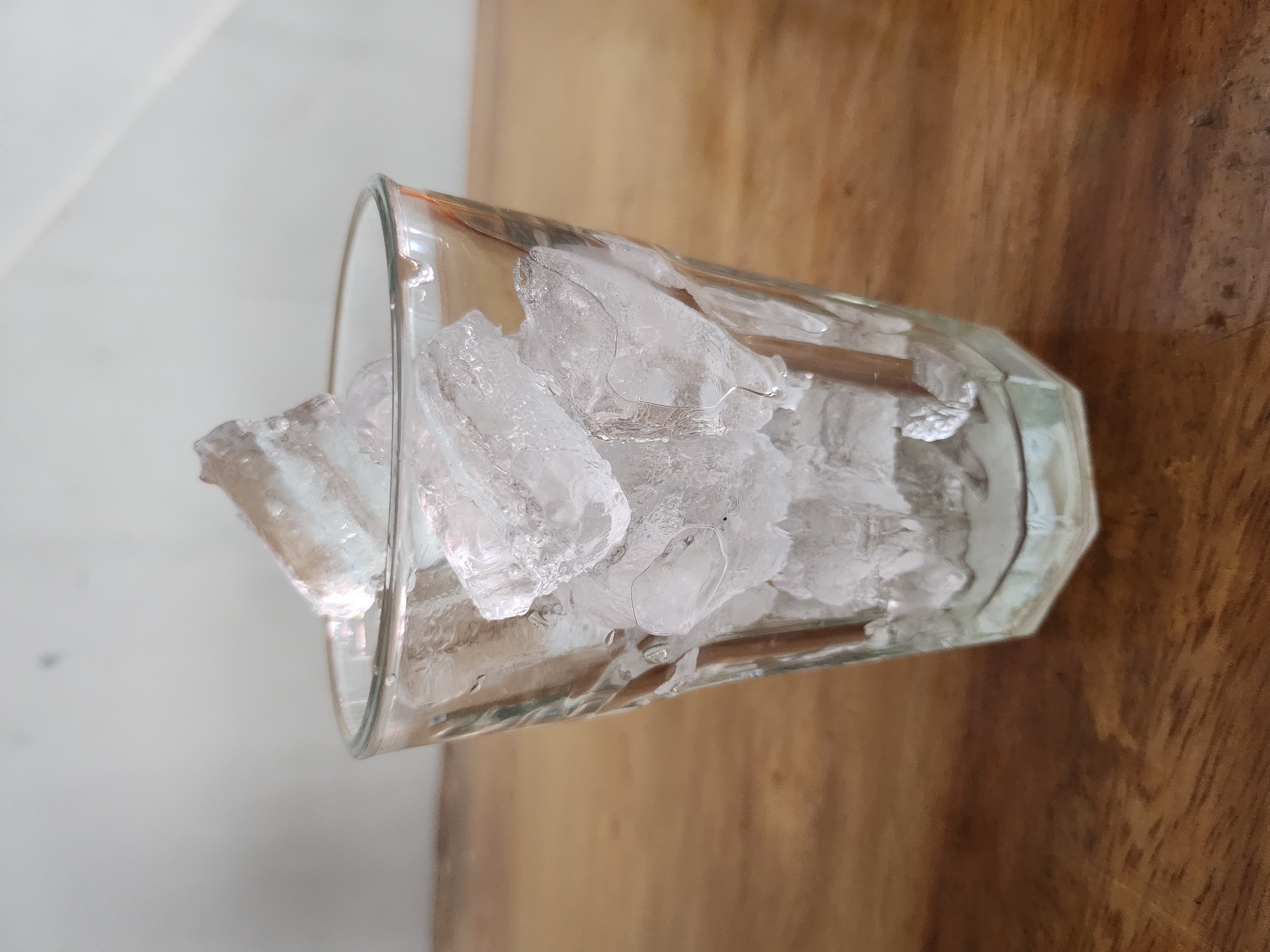 A close-up, eye-level shot captures a clear glass tumbler filled with crushed ice cubes. The glass, with a hexagonal base and vertical fluting, sits on a wooden surface, likely a table. The ice is piled high, with some pieces extending above the rim of the glass, creating a sense of abundance and coldness. The background is a plain, light-colored wall, with subtle shadows suggesting a light source from the right. The overall impression is one of simplicity and focus on the refreshing contents of the glass. The scene is devoid of human presence and is a static composition. The location in Depok, Indonesia, is not visually discernible from the image itself, as the scene lacks any culturally specific or geographically indicative elements.