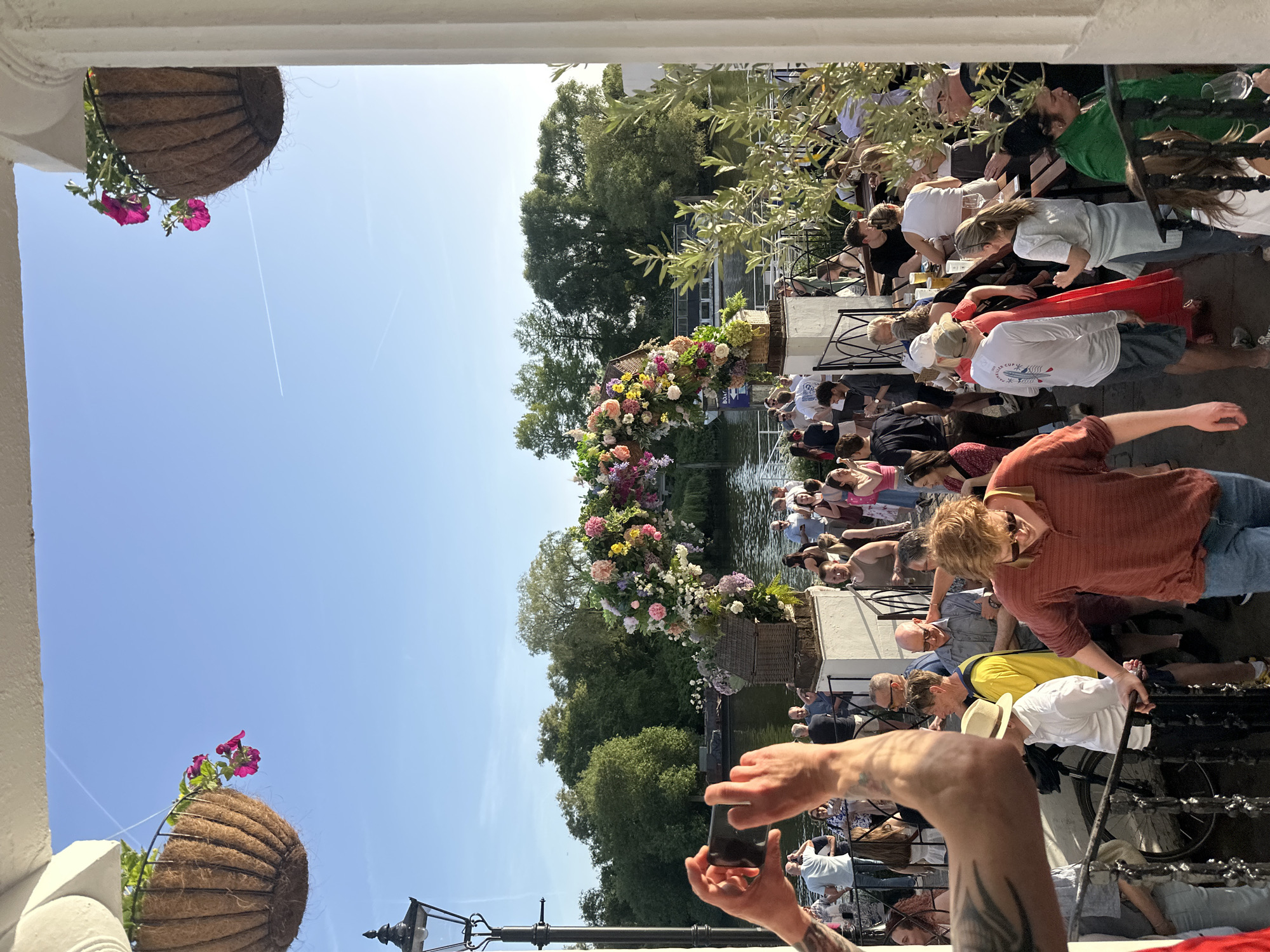 The image shows a scene on a canal in London, United Kingdom. A crowd of people is gathered on the side of the canal, with some seated at tables. There are hanging baskets with flowers decorating the scene, and the canal is lined with trees. In the foreground, a person is holding up a smartphone, presumably taking a photo. The environment appears to be outdoors on a sunny day. A lamppost stands in the foreground.