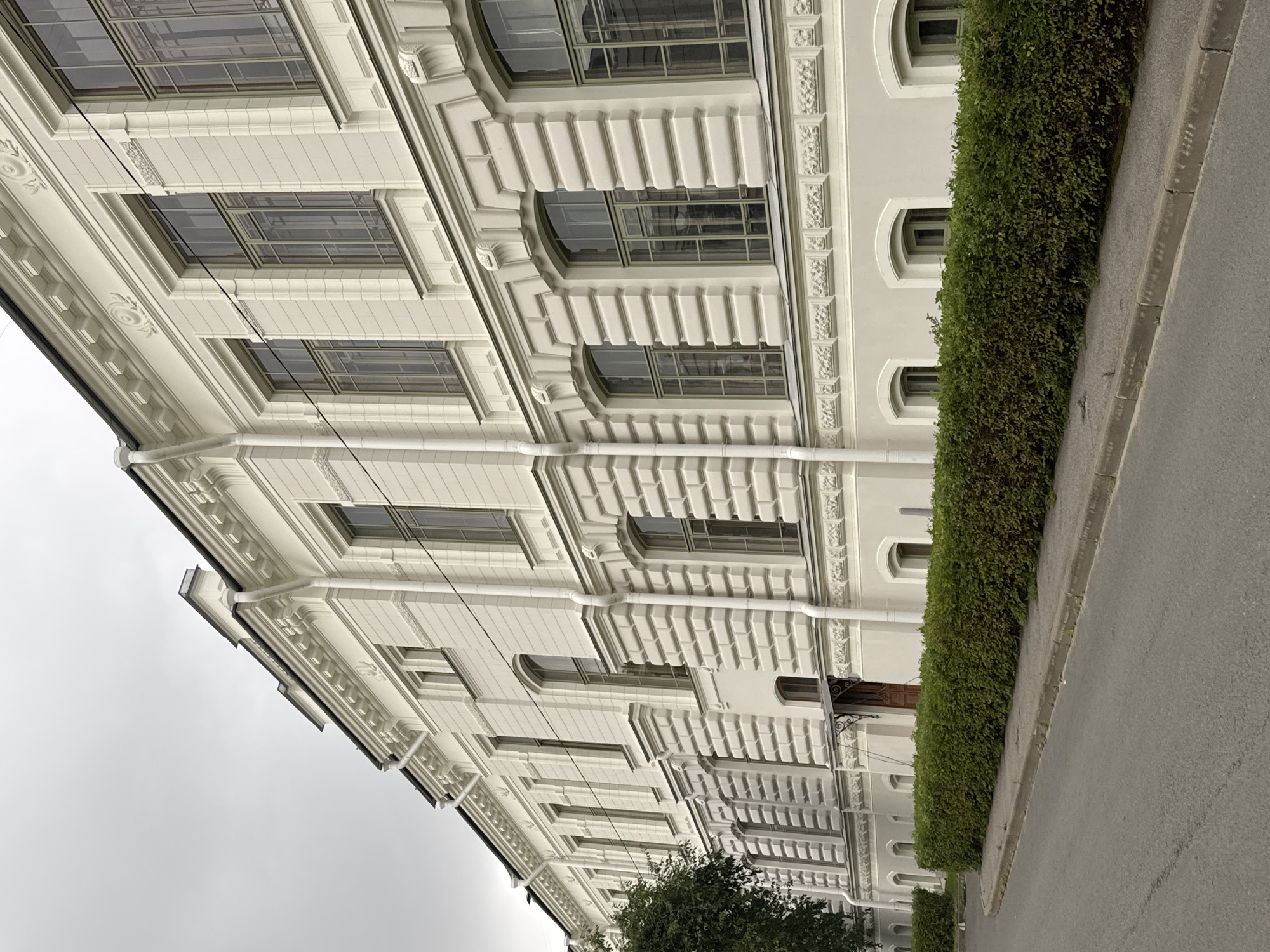 The image shows a side view of a light-colored, multi-story building with a classical architectural style. The building features numerous tall, rectangular windows with dark frames, many of which are divided into multiple panes. There are decorative architectural elements, including cornices, moldings, and recessed brickwork patterns on the facade. A green, neatly trimmed hedge runs along the front of the building, separating it from a paved sidewalk and an asphalt road. The sky is overcast and gray. The building appears to be located in a city, possibly Ladozhskoe Ozero, Russia, given the context, and it is a calm, overcast day with no visible people or activity.