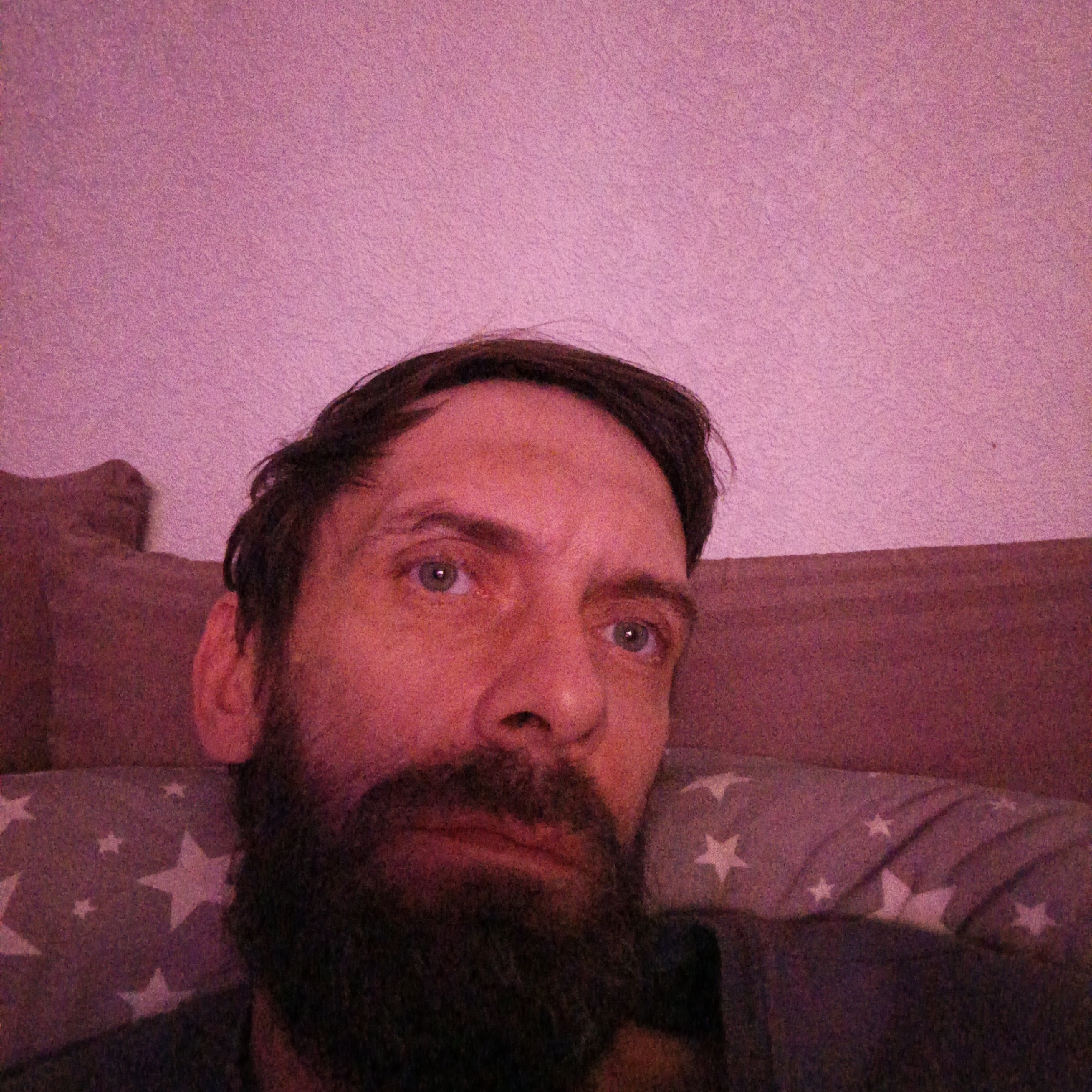 A close-up selfie depicts a middle-aged man with dark hair, a beard, and blue eyes. He is lying down, with his head resting on a pillow patterned with white stars. The background consists of a pink wall and what appears to be the headboard of a bed or a pillow. The location in the city of Марковский, Russia is not discernible from this image.