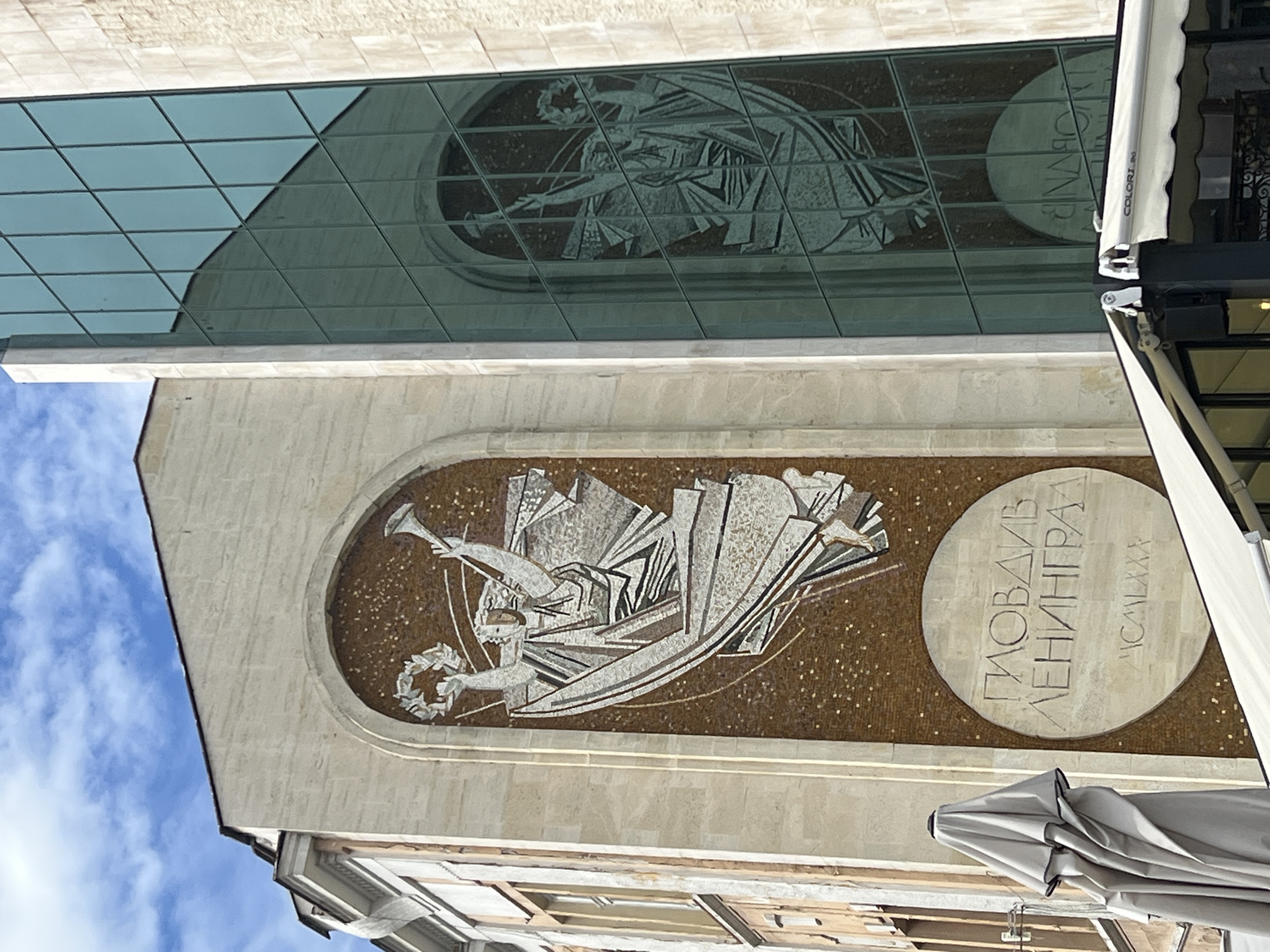 The image shows a mosaic on a building in Plovdiv, Bulgaria. The mosaic depicts a stylized figure of a person with a trumpet and a wreath. Next to the figure is a round panel with Cyrillic text, "Пловдив Ленинград", and the Roman numerals "MCMLXXX". The mosaic is set against a background of small brown tiles. Above the mosaic is a modern glass facade, which reflects a similar mosaic. Parts of awnings are visible on the right side of the image and a blue sky with clouds are visible to the left.