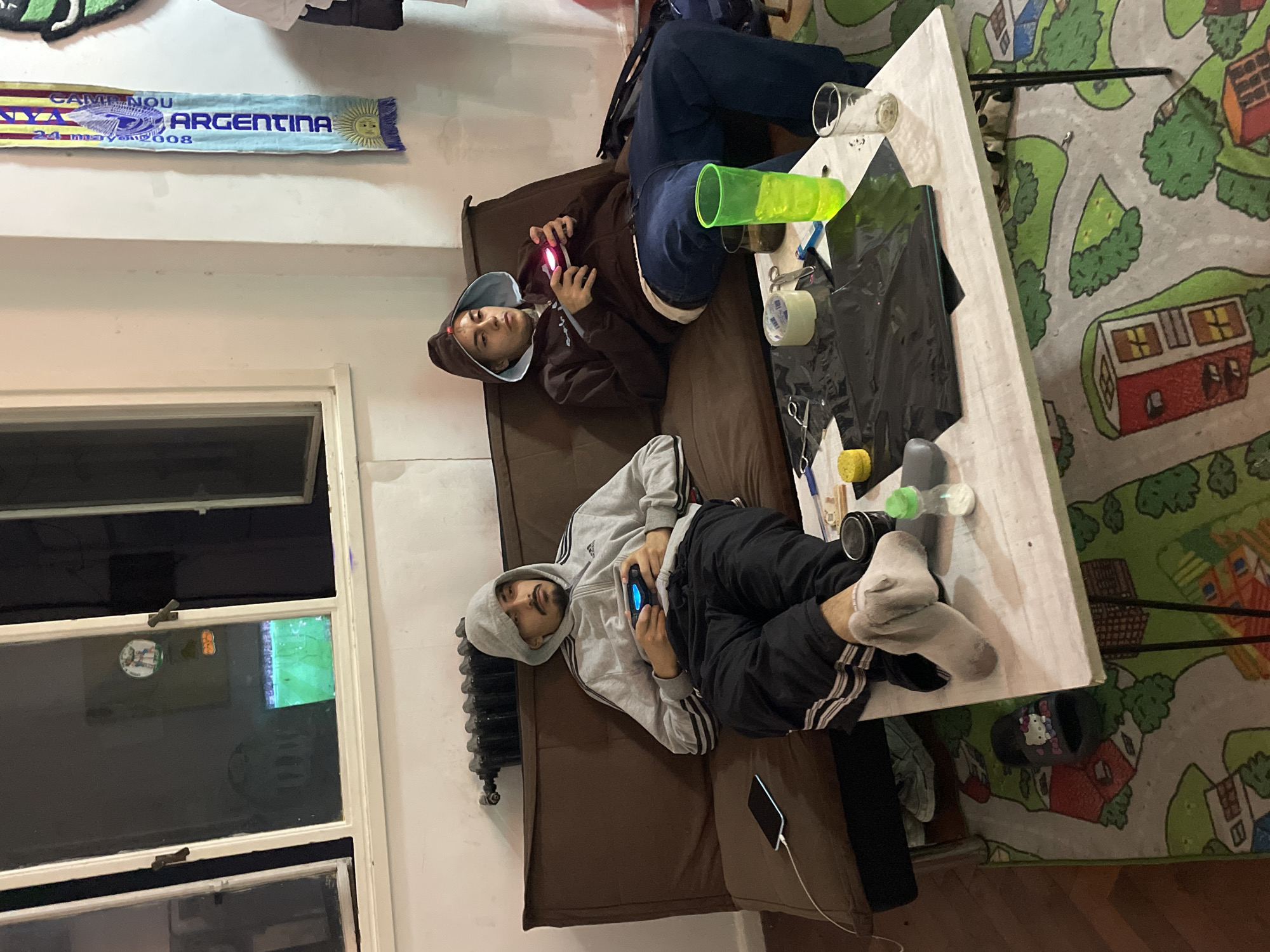 Two males are lying on a brown couch. One male is wearing a brown hooded sweatshirt, while the other male is wearing a gray hooded sweatshirt. Both males are holding video game controllers. A white table is positioned to the right of the couch. The table has various objects on top of it, including a clear glass, a green plastic container, a roll of tape, scissors, and black plastic bags. A children’s rug with houses and trees is under the table. A window is visible to the left of the couch. Above the couch, a scarf with the text "Camp Nou Argentina 24 Mayo 2008" is hanging. A radiator is next to the couch. A phone is connected to a charging cable. Based on the scarf displaying "Argentina," the location is likely in Buenos Aires, Argentina.