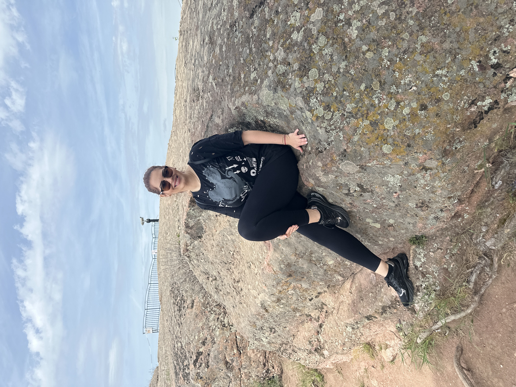 A woman in sunglasses, a dark t-shirt, and dark leggings is seated on a large rock formation. Her left hand is touching the surface of the rock behind her. The rock is textured with lichen growth. In the background, there is a view of a blue sky with white clouds and a distant metal fence structure. The ground near the rock base appears reddish. The photo was taken at Belogradchik, Bulgaria.
