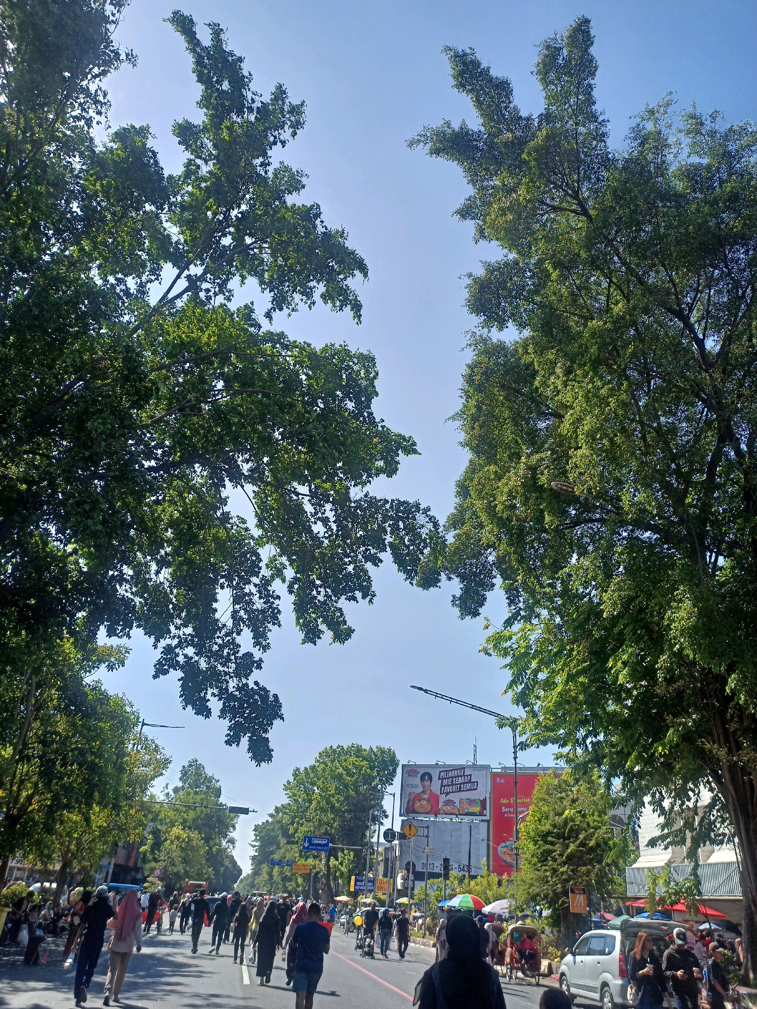 A busy street scene unfolds in Surakarta, Indonesia, under a clear blue sky. The foreground is dominated by the lush green canopy of trees lining the street, casting dappled shadows. A moderate number of people are walking along the road, some in groups, others individually. Many are wearing casual clothing, and some women are seen wearing headscarves. Several vehicles, including a silver minivan, are present on the road, along with a rickshaw.
In the background, large billboards are visible, advertising various products and services. Traffic signals and street lights indicate an urban setting. The overall impression is one of a vibrant public space filled with activity on a sunny day. The street appears to be a main thoroughfare, likely a place where commerce and daily life intersect. The presence of trees suggests a well-maintained urban environment. The scene is dynamic, with people moving and interacting within the bustling city.