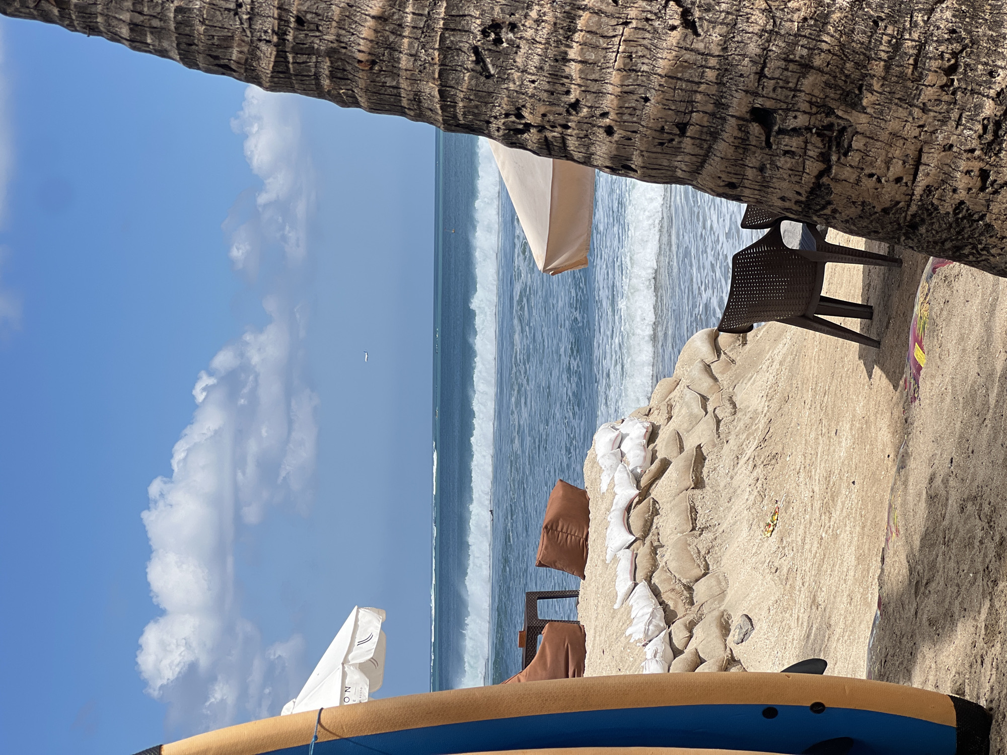 A bright, sunny day at a beach in Kuta, Indonesia, is depicted, with a focus on leisure and surf. In the foreground, a surfboard, painted with blue and yellow stripes, stands vertically on the sand, leaning slightly to the left. To the right, the textured trunk of a palm tree dominates the frame, obscuring a portion of the view.
The middle ground reveals a sandy beach area with several pieces of outdoor furniture arranged on a raised sandbag embankment. Two brown beanbag chairs and a small dark side table are positioned under a white umbrella with visible text. Further back, another umbrella, light tan in color, is partially visible. Two dark brown plastic chairs are set up closer to the water's edge, facing the ocean.
The ocean itself is a vibrant blue with moderate waves breaking. Several surfers are visible in the distance, riding the waves. The sky above is a clear, bright blue, dotted with fluffy white clouds. An airplane is tiny and visible in the far distance. The scene suggests a relaxed atmosphere, perfect for enjoying the surf and sun.