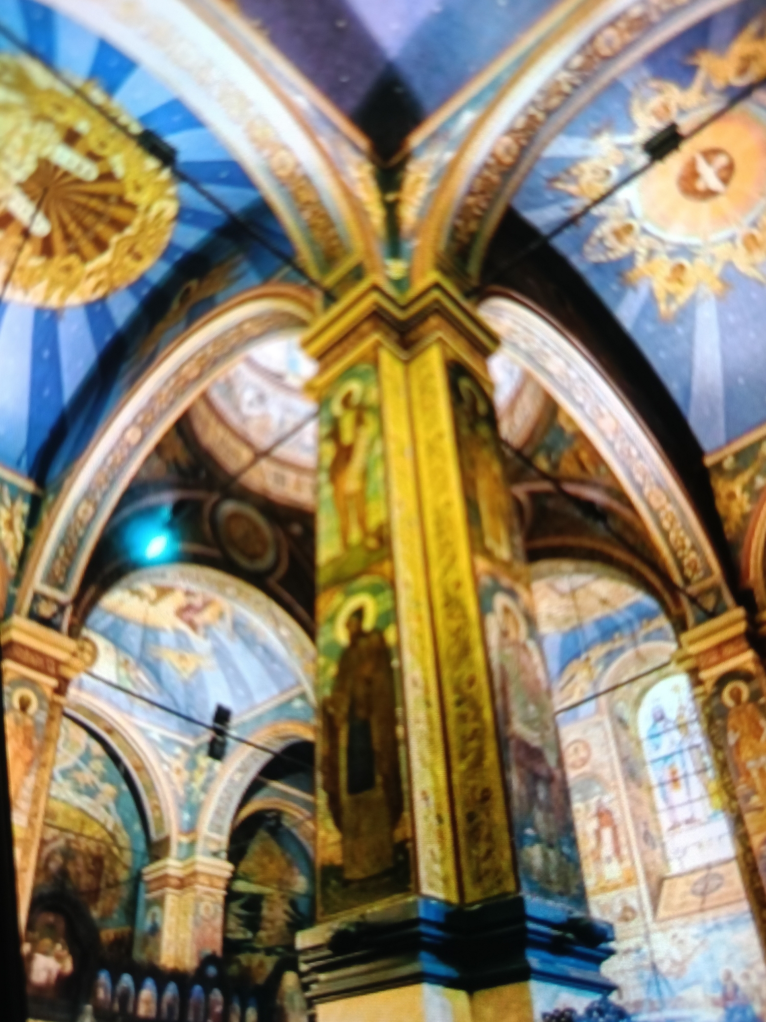 This is an interior view of a church or cathedral in Varna, Bulgaria. The image captures the ornate ceiling and a large central pillar, both extensively decorated with vibrant frescoes. The ceiling is a deep blue, adorned with radiating golden patterns and depictions of figures, possibly angels or saints. Arches with golden trim support the ceiling structure. The central pillar is also covered in detailed paintings, featuring figures in religious attire. Other architectural elements, like smaller arches and sections of walls, are visible in the background, also covered in frescoes depicting biblical scenes or figures. A stained-glass window is visible to the right. The overall impression is one of grandeur and religious devotion. The lighting suggests an interior setting, possibly during daytime, with artificial lighting contributing to the illumination. There is no visible text in the image.
